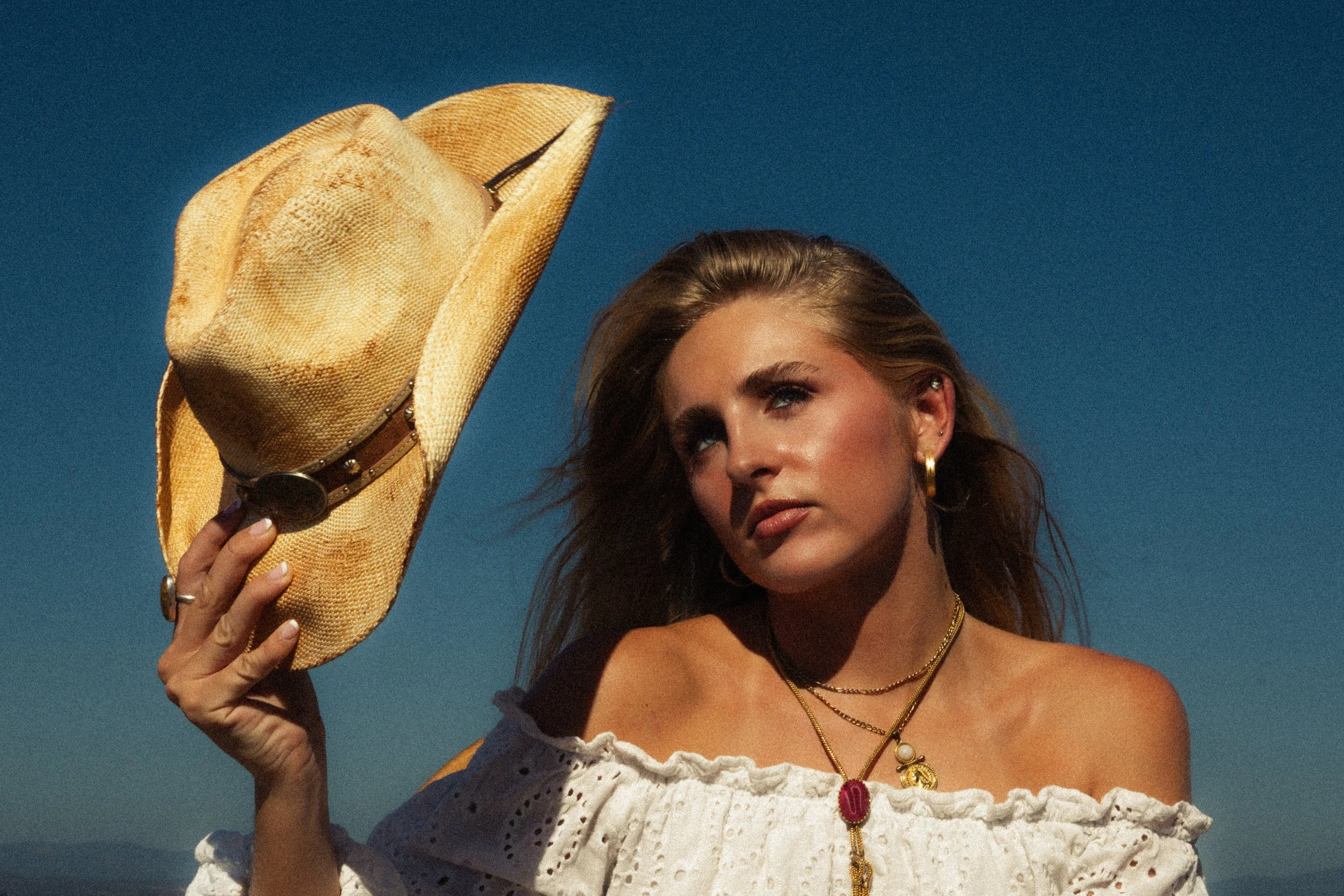 Woman with brown hair, gold jewelry, and wearing a white lace top, holding a straw hat with her right hand.