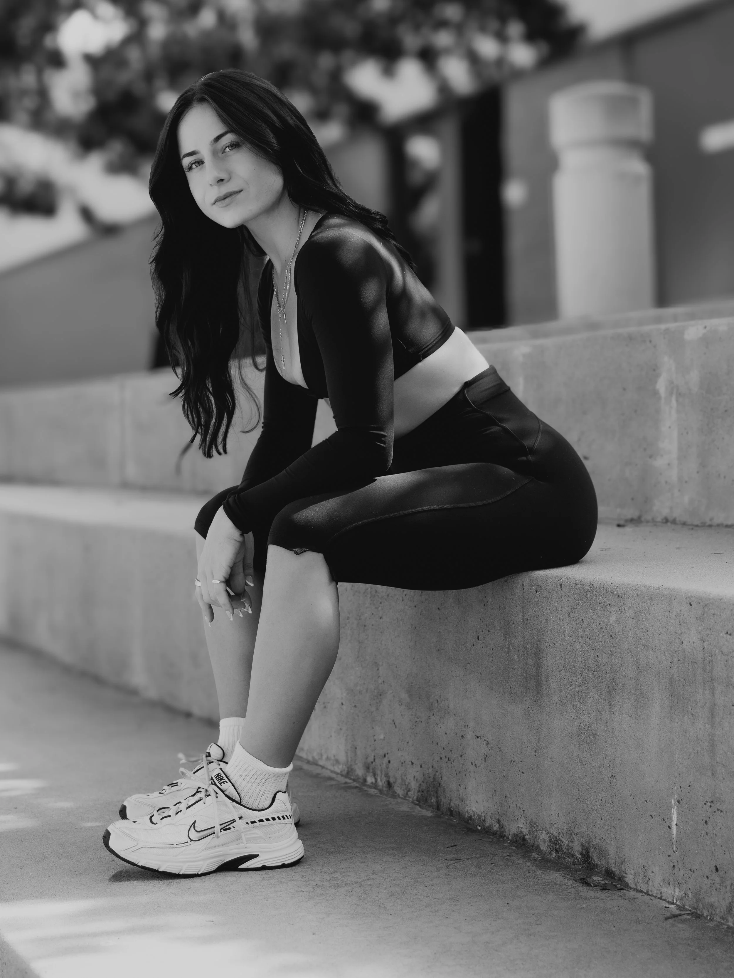 A young woman with long dark hair sitting on a concrete ledge outdoors, wearing athletic clothing and sneakers, with a blurred natural background.