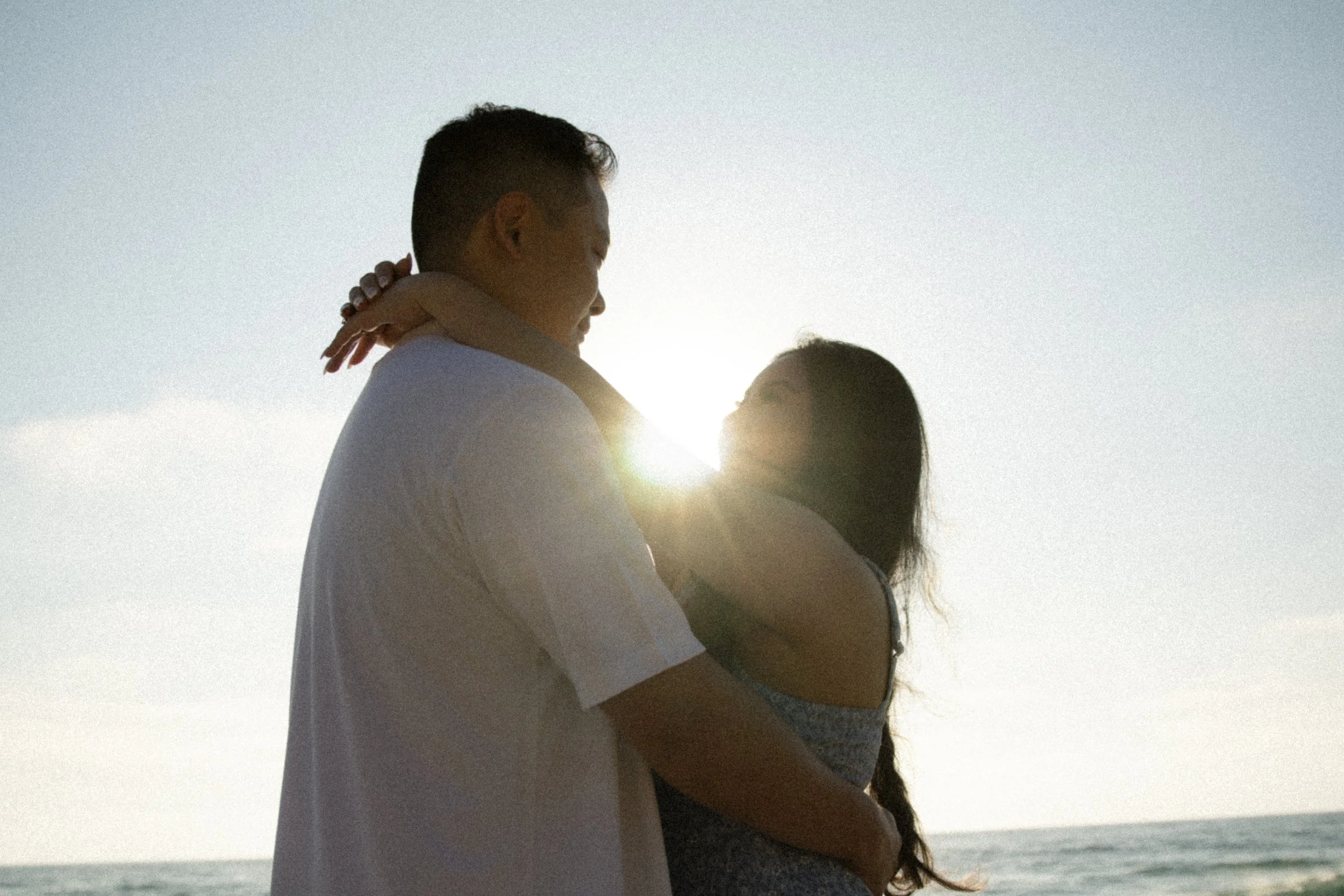 A couple embraces on the beach at sunset, with the ocean in the background and the sun setting behind them.