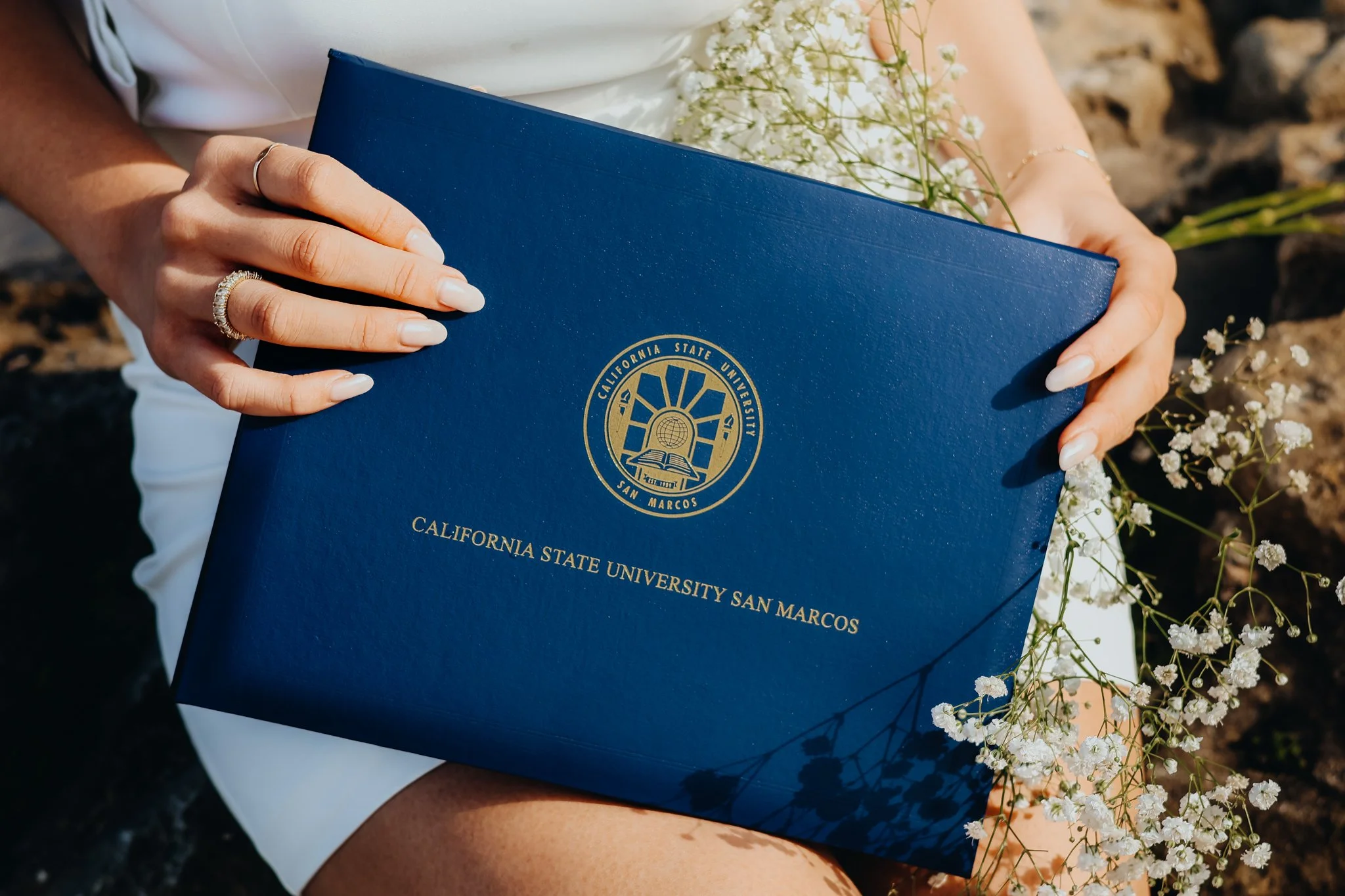 A person holding a blue California State University San Marcos diploma cover, with white baby’s breath flowers nearby.