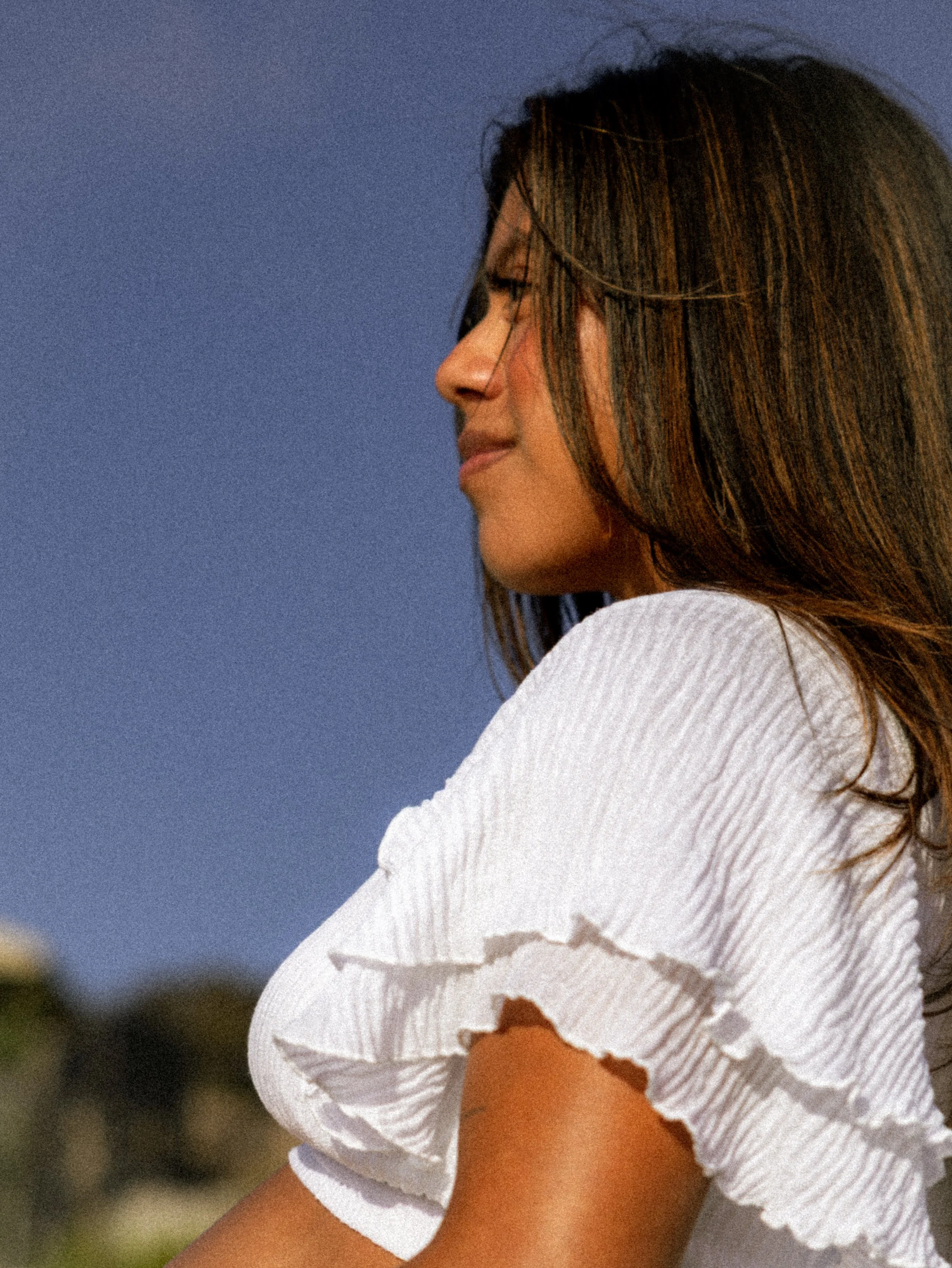 A woman with long, brown hair and a light complexion, wearing a white, ripped short-sleeved shirt, is smiling with her eyes closed against a blue sky background.