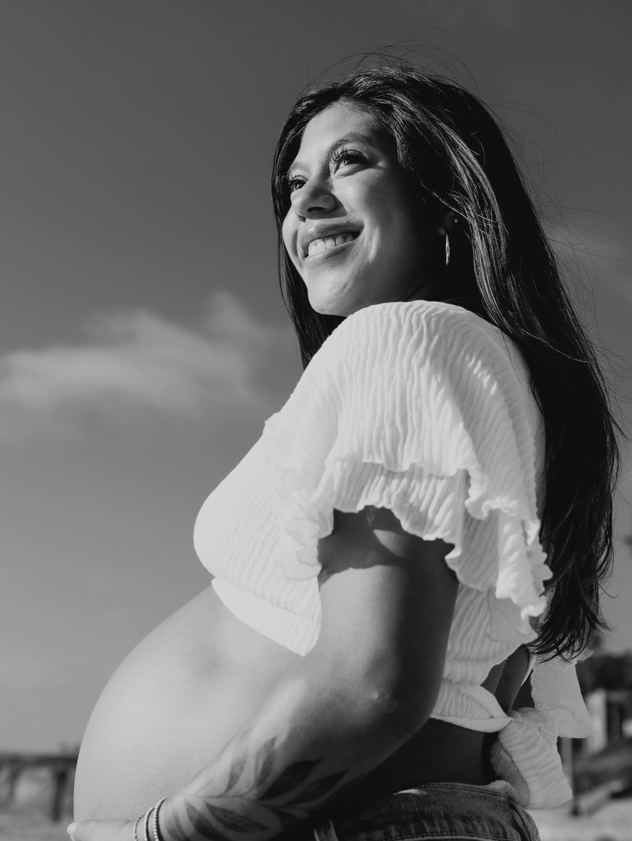 Black and white photo of a woman smiling, looking up with wind blowing her hair, wearing a light-colored top with ruffled sleeves and earrings, outdoor background.