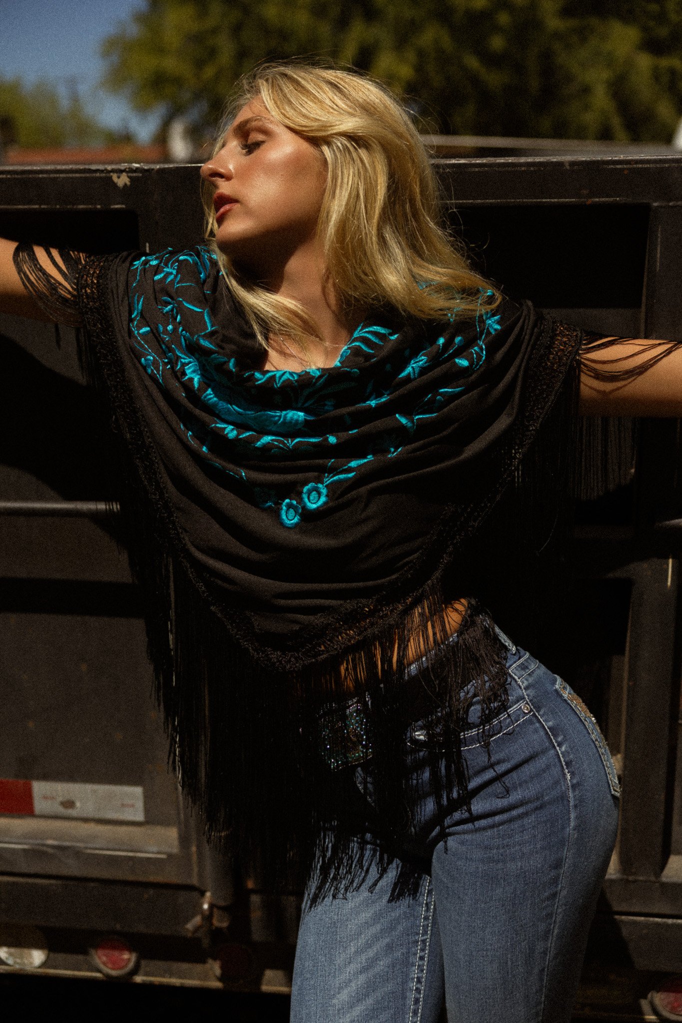 A woman with long blonde hair posing outdoors at night, wearing a black top with blue embroidered details and blue jeans.