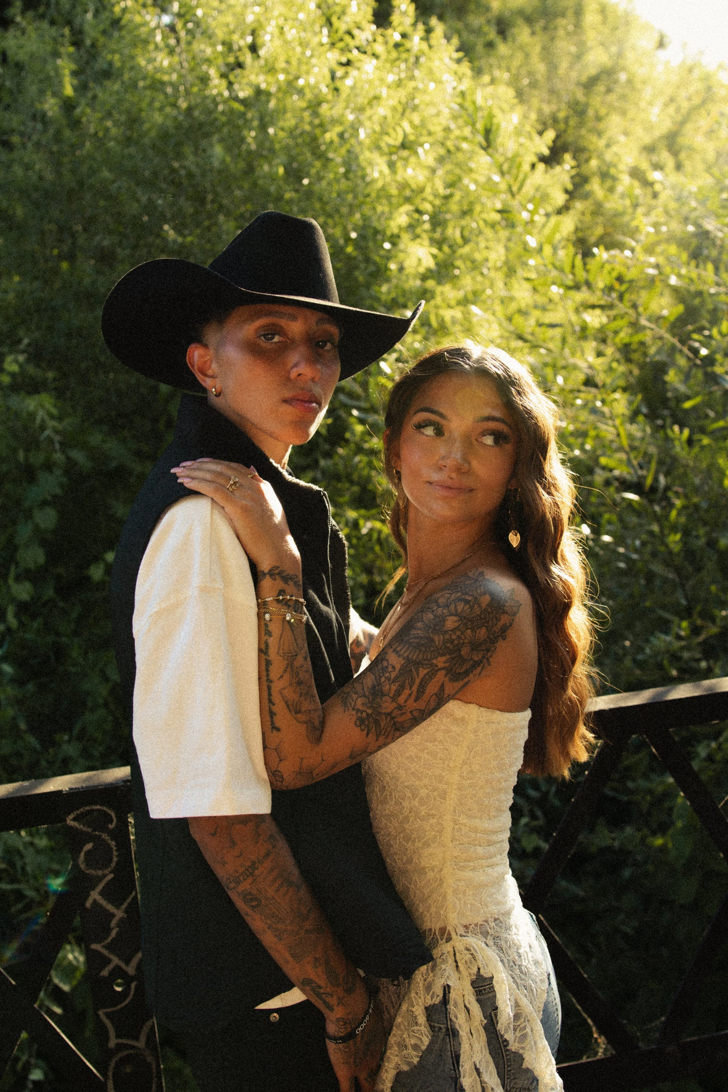 Two women standing close together outdoors in sunlight, with lush green foliage in the background. One woman has short dark hair, a black wide-brimmed hat, and tattoos on her arms. The other woman has long wavy hair, tattoos on her arm, and is wearin
