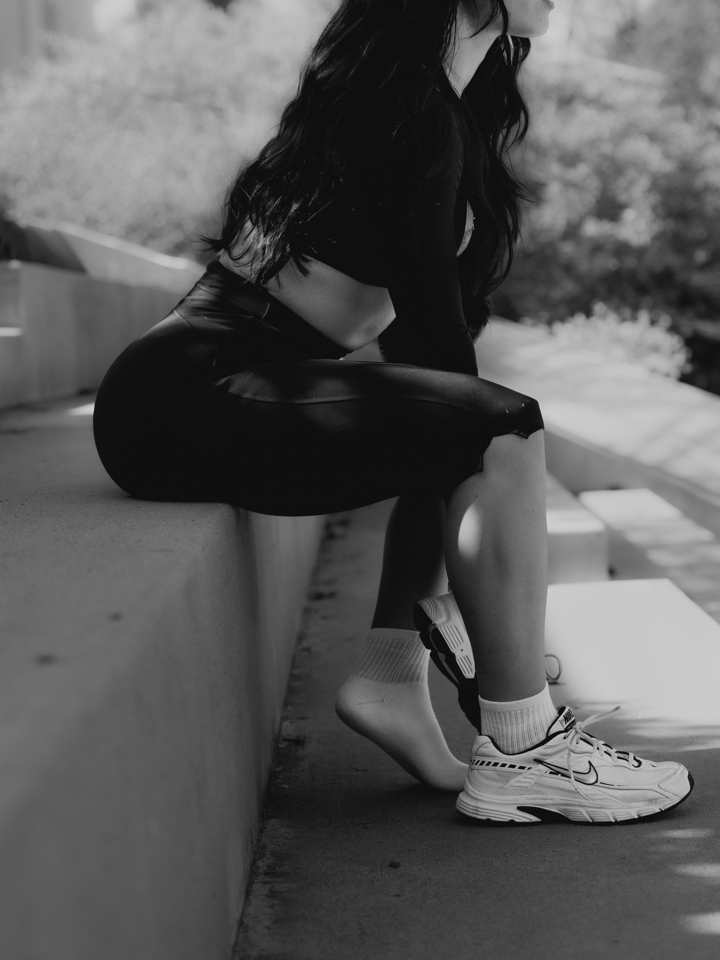 A person with long dark hair, dressed in athletic clothing, sitting outdoors on a concrete ledge, wearing running shoes and socks, with a blurred background of trees and natural surroundings in black and white.
