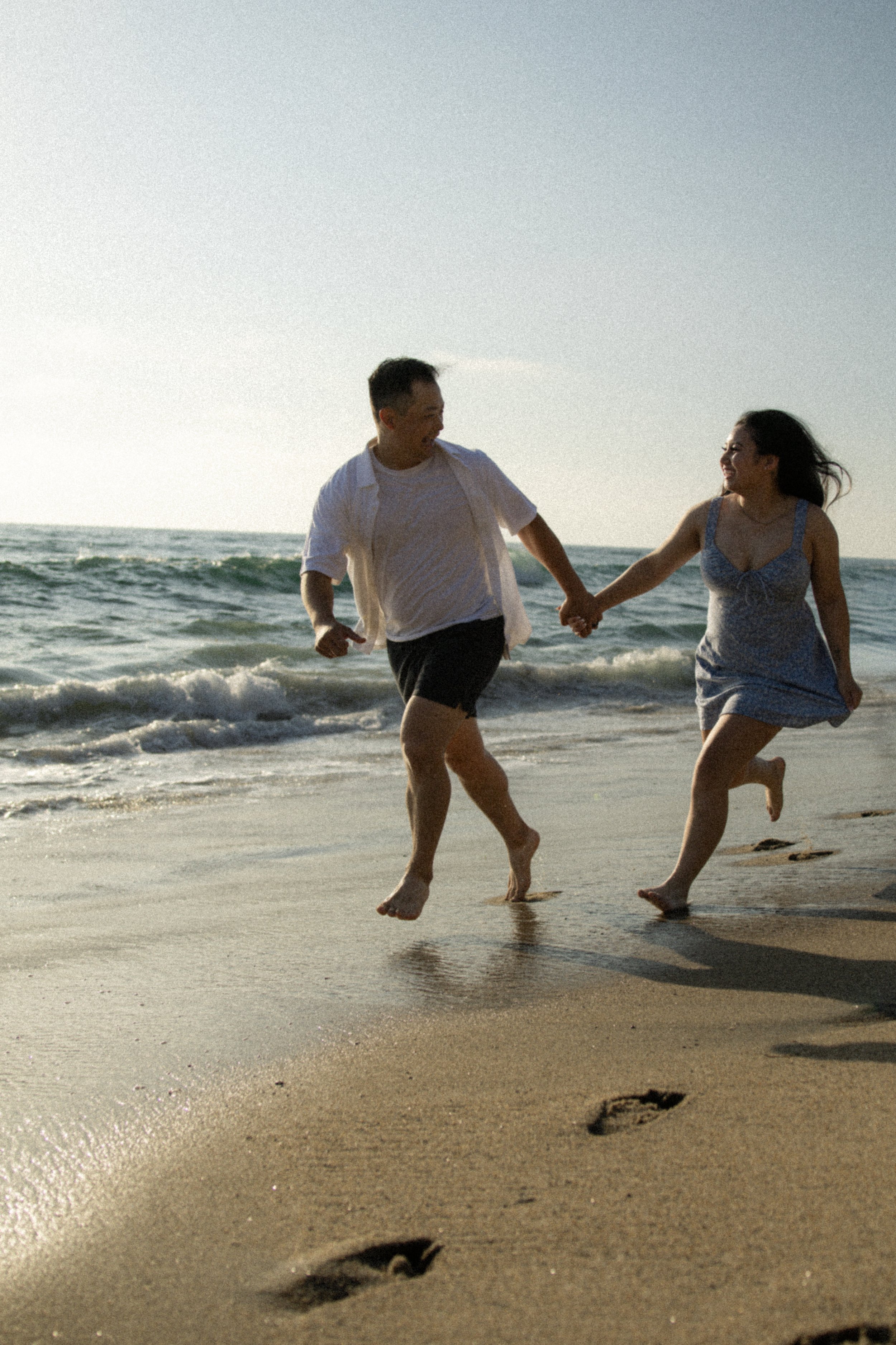 A man and woman running hand in hand along the shoreline of a beach, with footprints visible in the sand and waves crashing behind them, during what appears to be late afternoon or early evening.