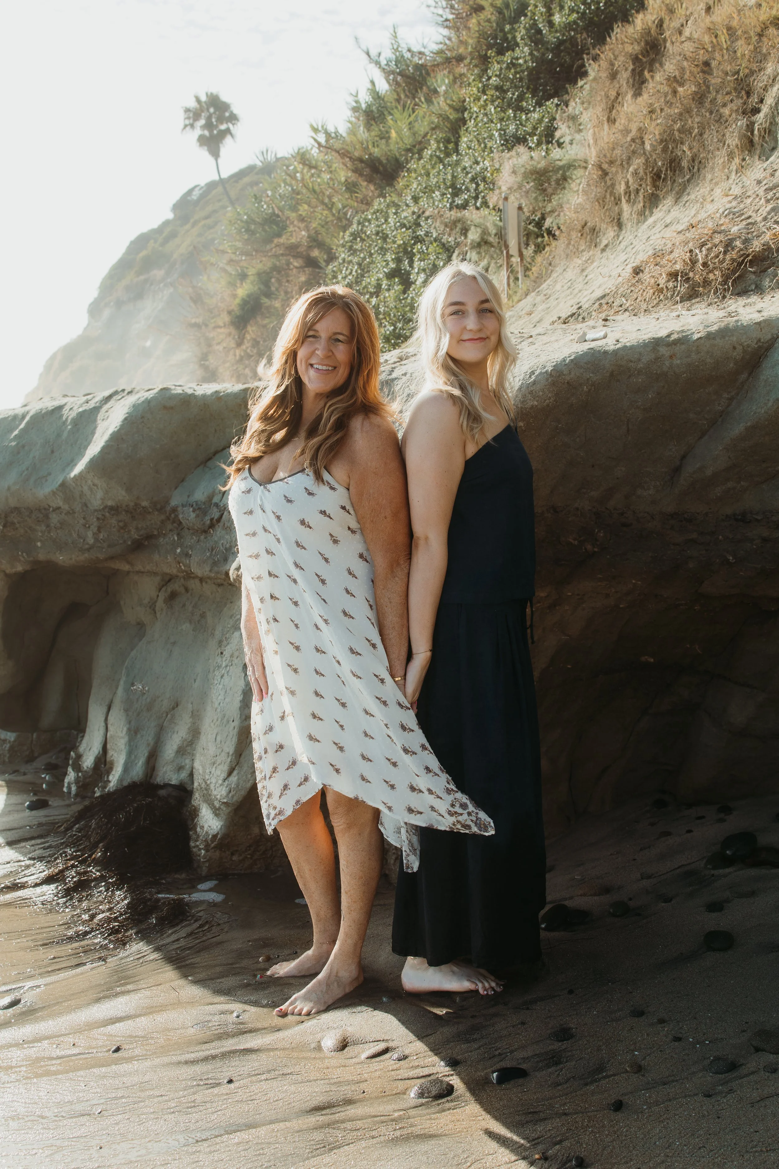 Two women standing barefoot on a sandy beach near rocks, with a hillside covered in greenery and trees behind them. The woman on the left has long, wavy auburn hair and is wearing a sleeveless white dress with a leaf pattern. The woman on the right h
