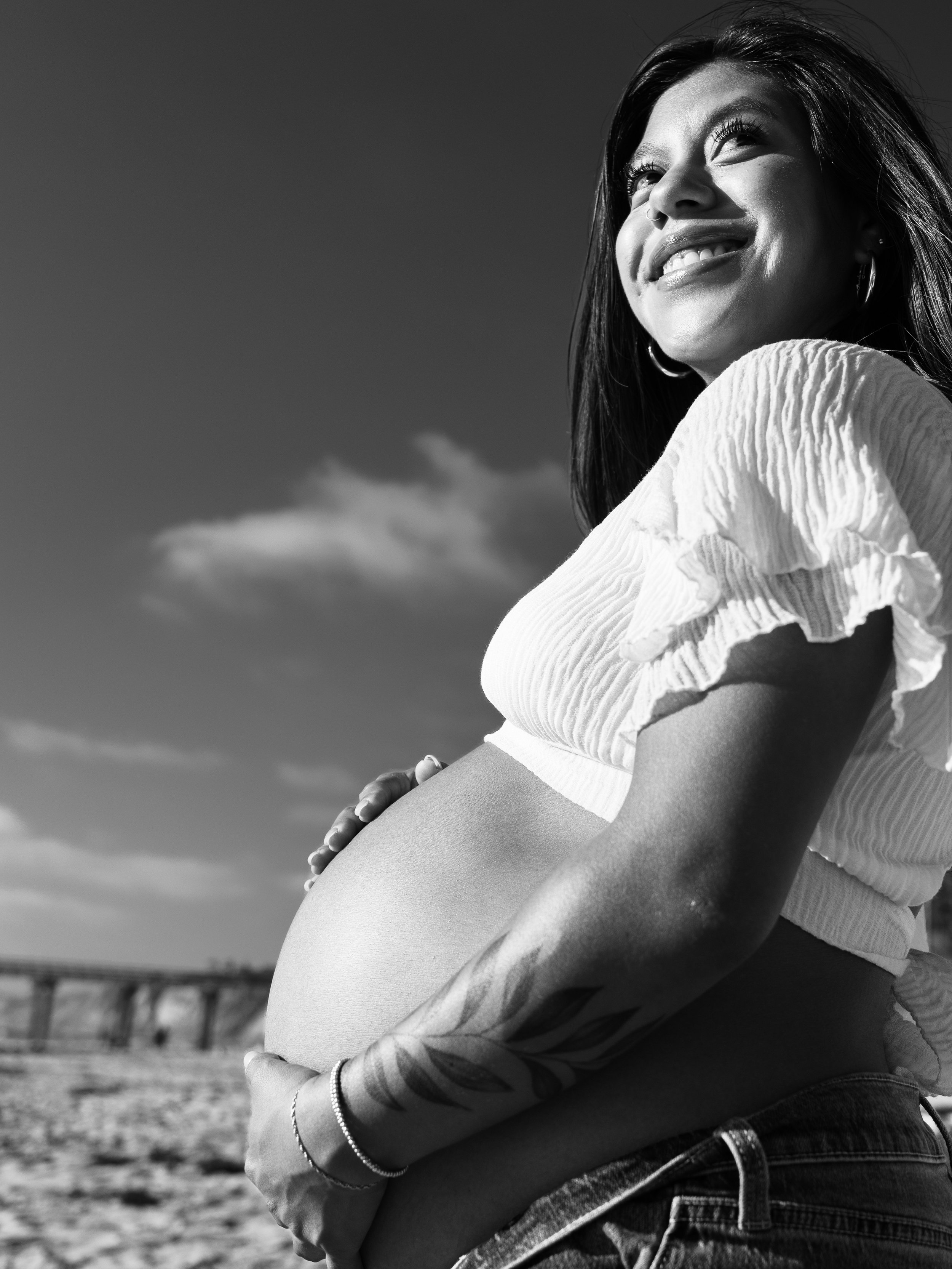 A young woman with dark hair and earrings, smiling and looking into the distance, with her arms crossed over her pregnant belly, outdoors with a cloudy sky in the background.
