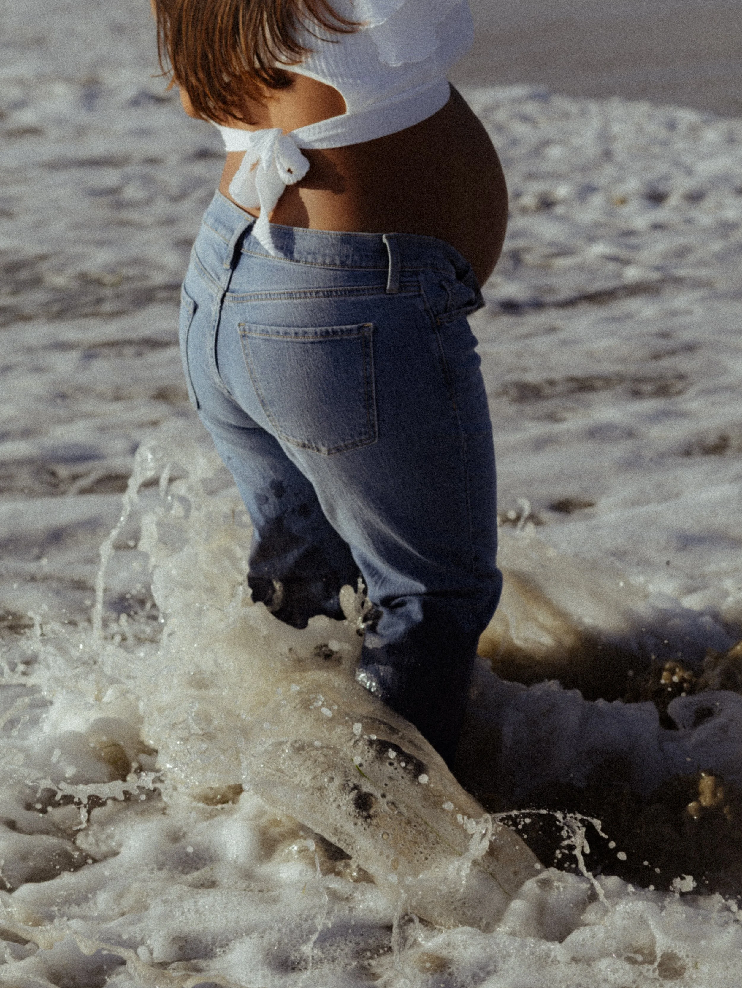 A pregnant woman wearing jeans and a tied white shirt on the beach, walking through the foamy ocean waves.