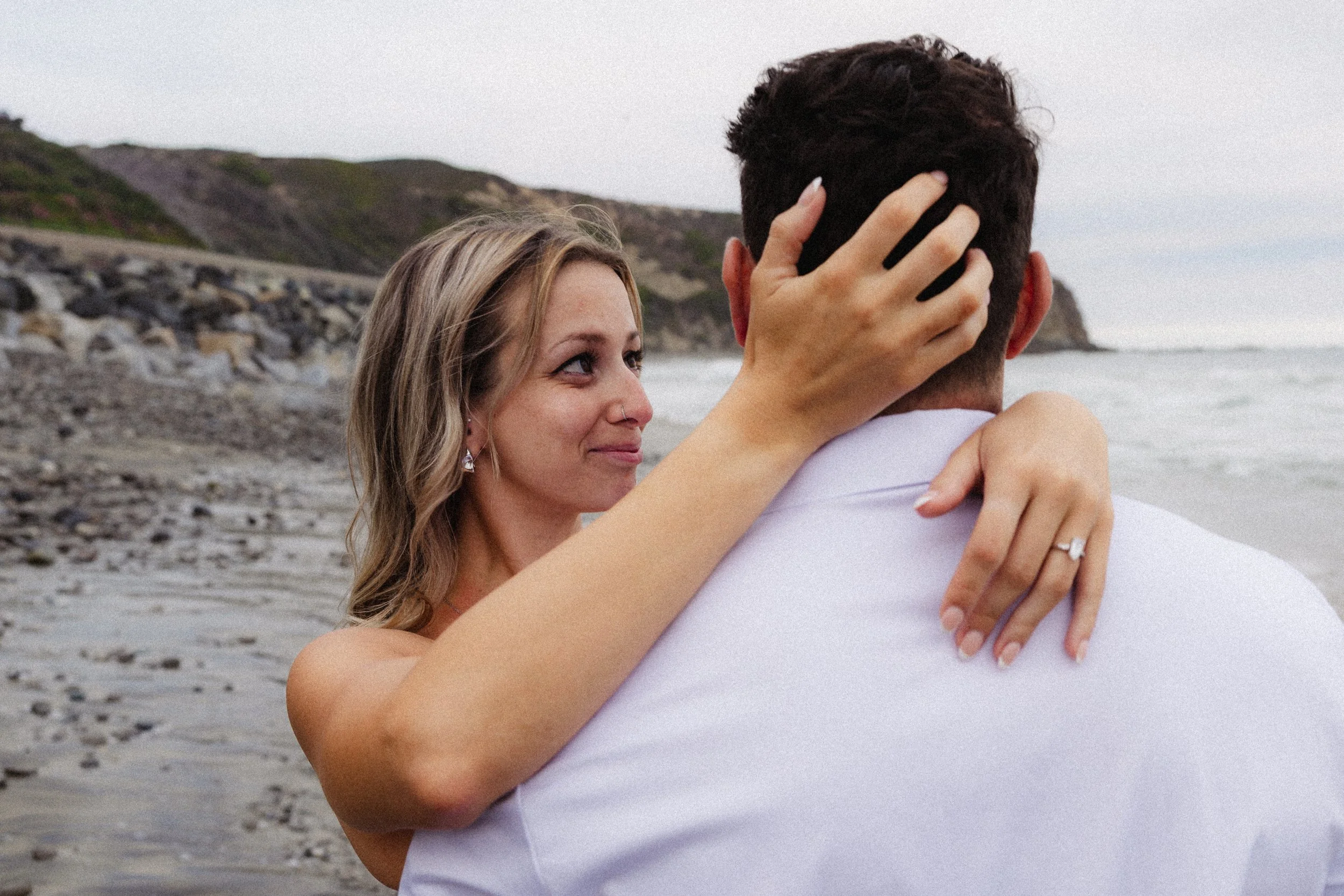 A woman with blonde hair and earrings is hugging a man on a beach. Her hand is on the back of his head, and she is smiling as she looks at him. The man has dark hair and is facing away from the camera. The background shows rocks, hills, and the ocean