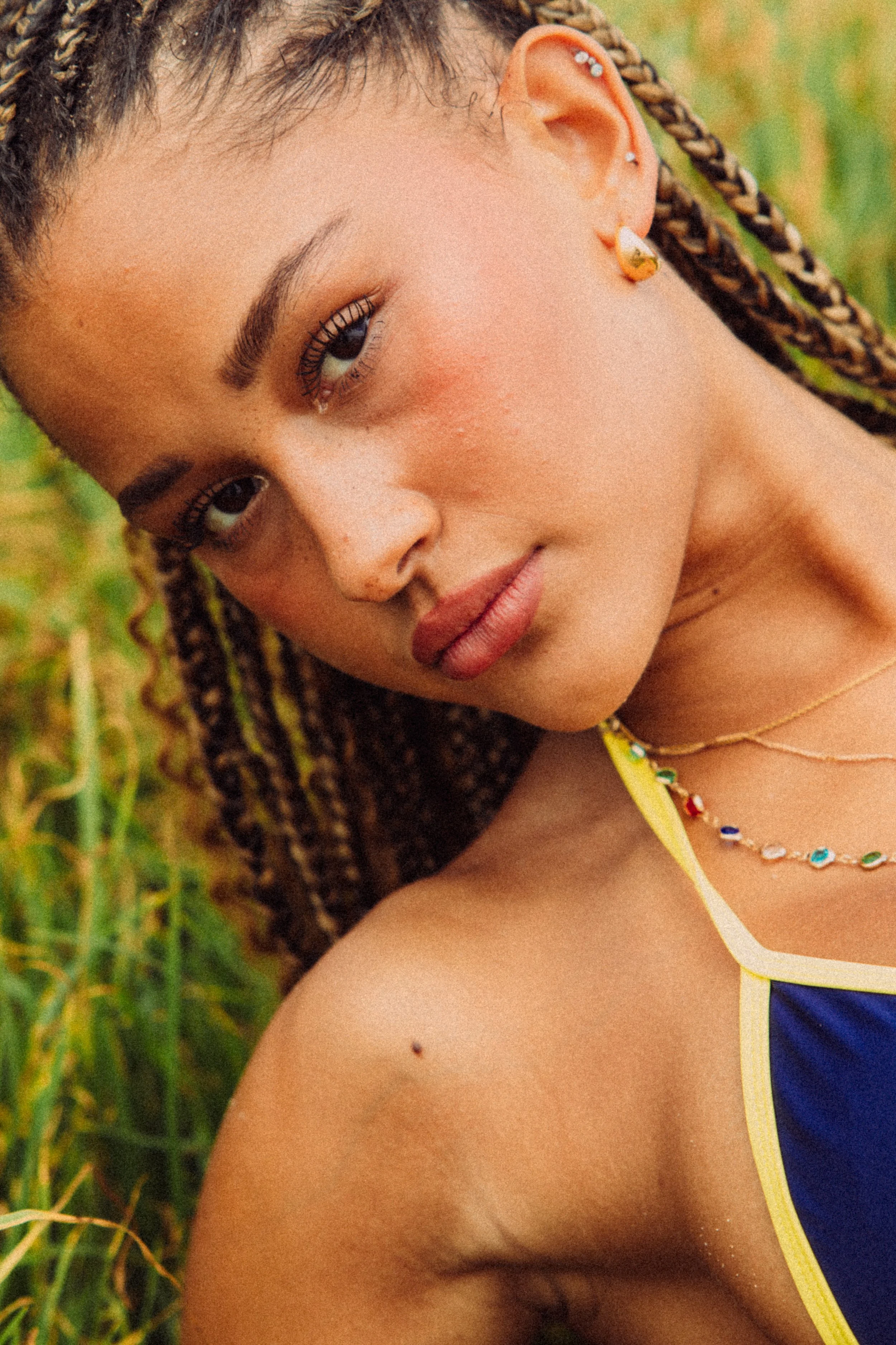 Close-up of a young woman with braided hair, earrings, and a beaded necklace, lying outdoors in a grassy area.