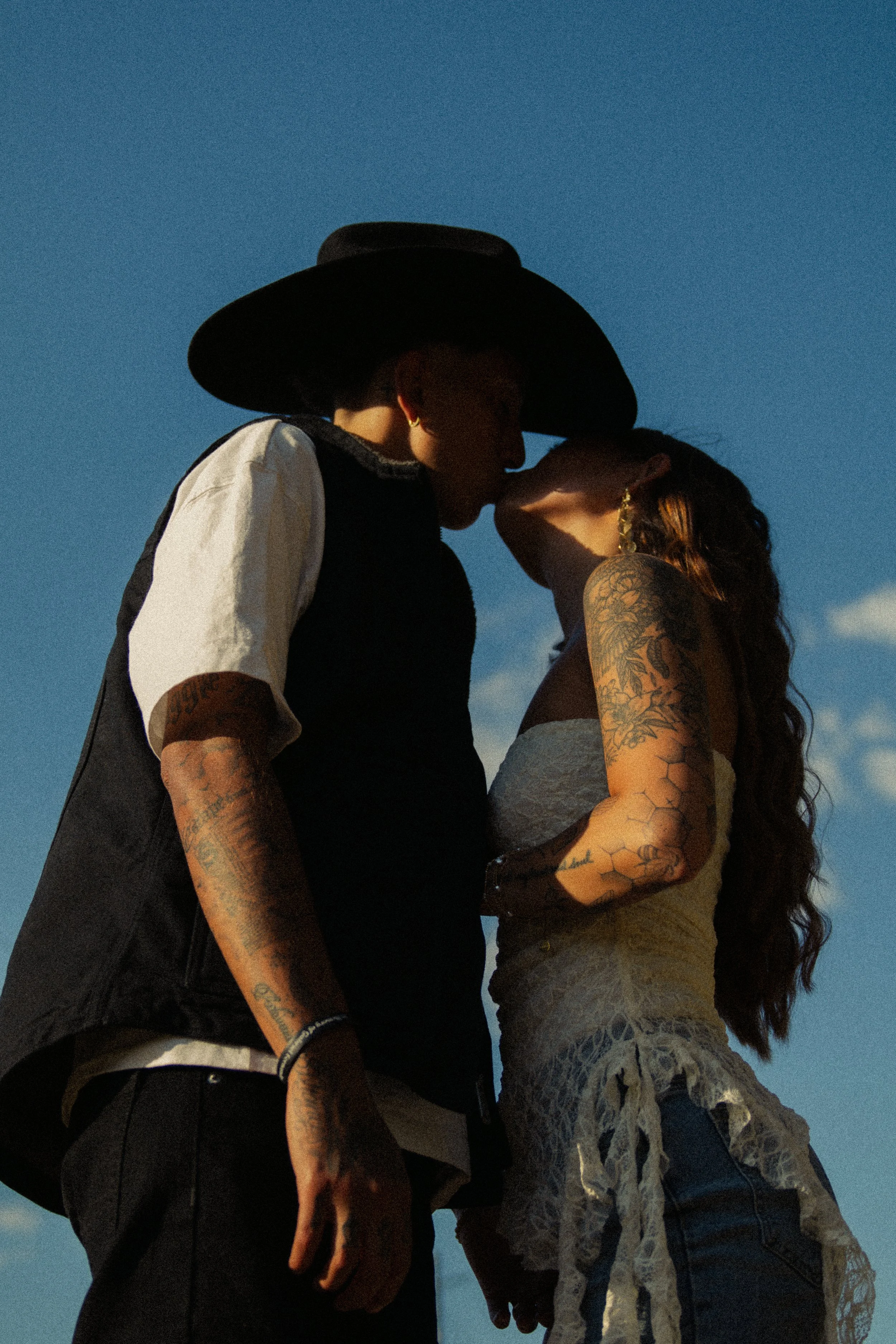 A man and woman standing close together outdoors against a blue sky, nose to nose in a tender moment, with their eyes closed.