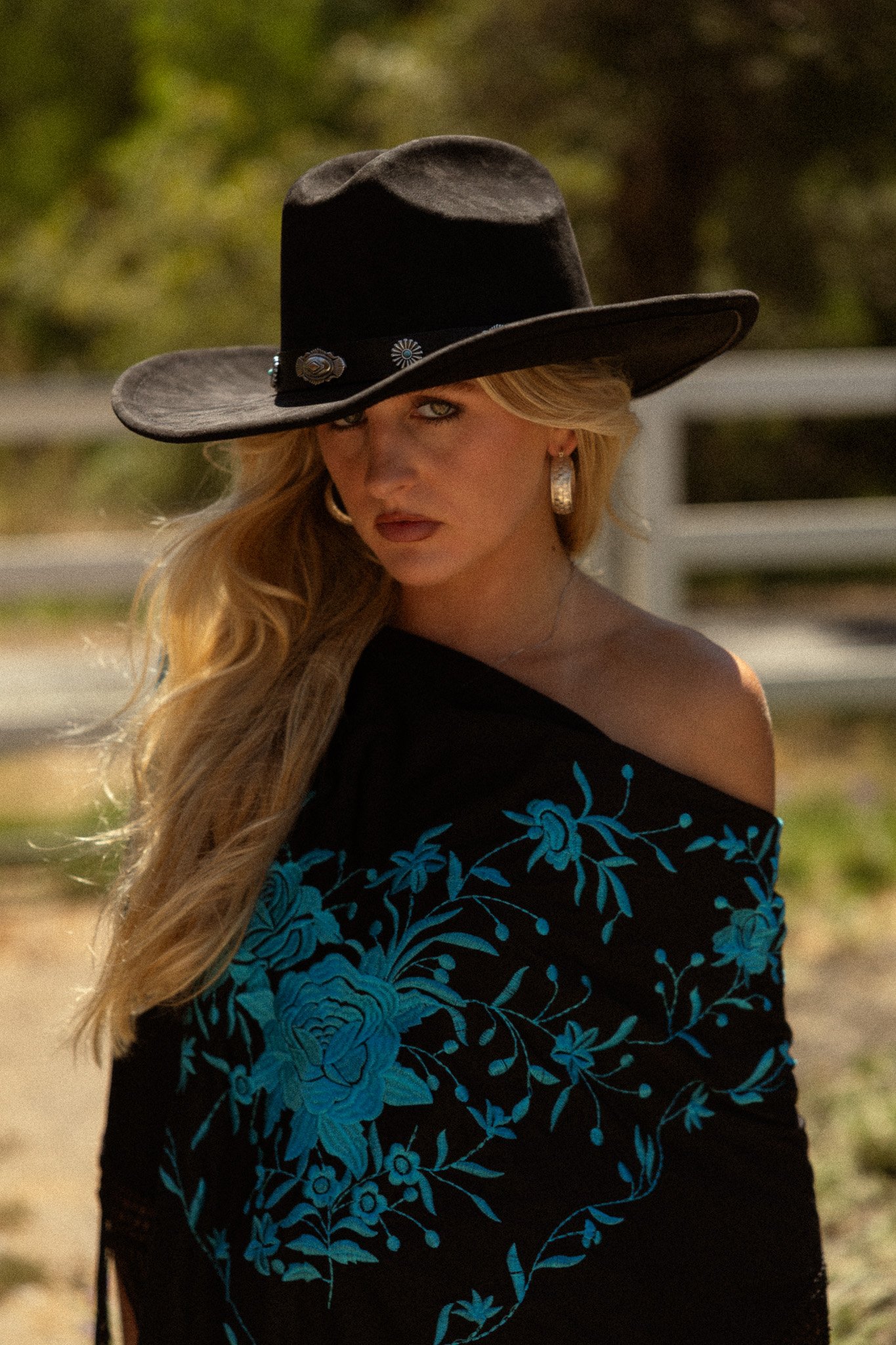 A woman wearing a large black cowboy hat with decorative silver conchos, gold earrings, and a black dress with blue floral embroidery.