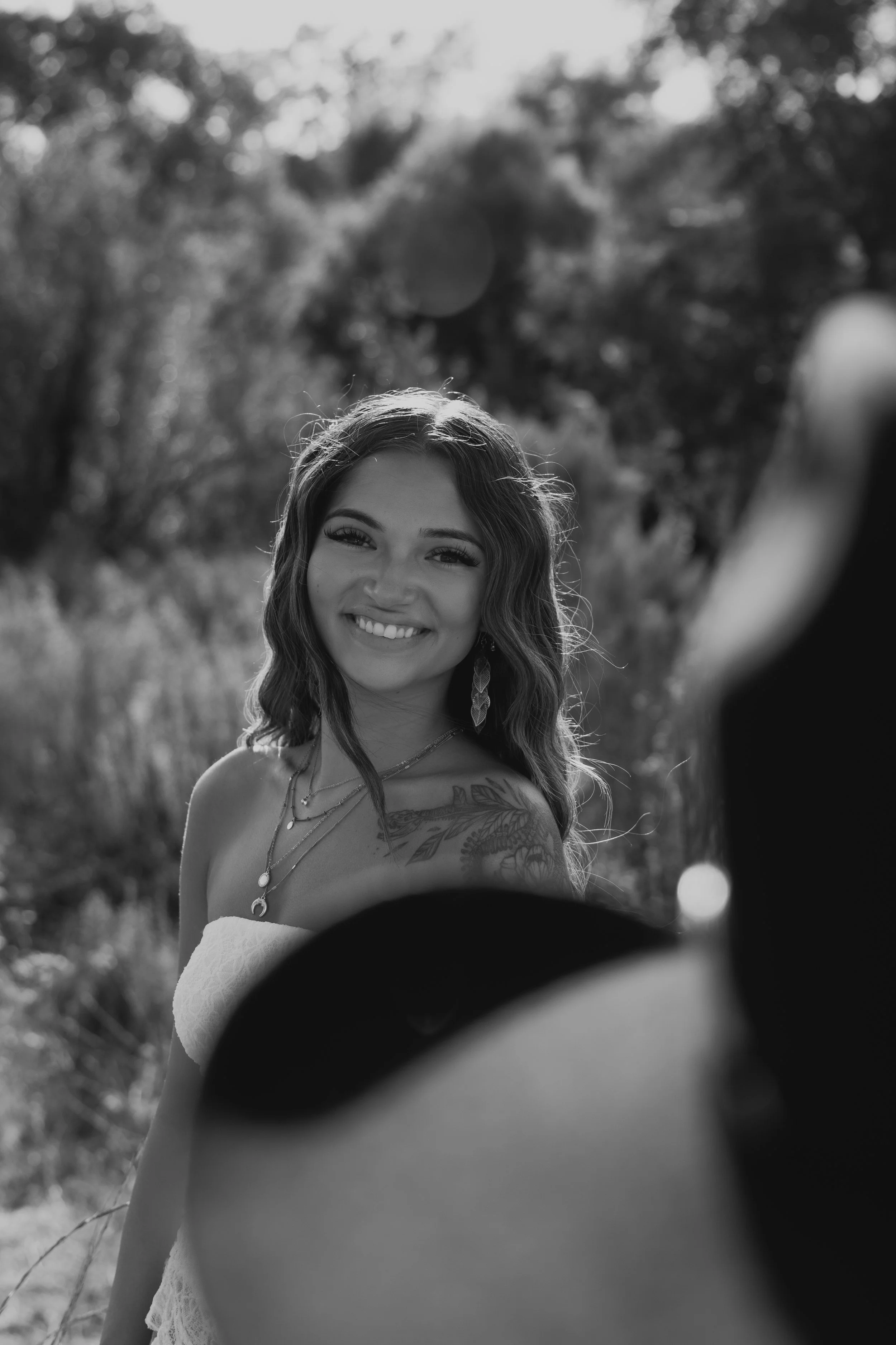 Black and white photo of a smiling young woman with long hair, wearing jewelry and a strapless top, outdoors with blurred trees and foliage in the background, partially framed by an out-of-focus dark circular object in the foreground.