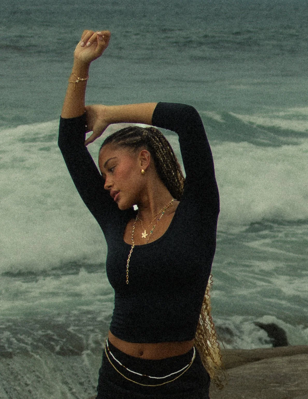 A woman with braided hair in a black long-sleeve top standing at the beach with ocean waves in the background, with her eyes closed and arms raised above her head.