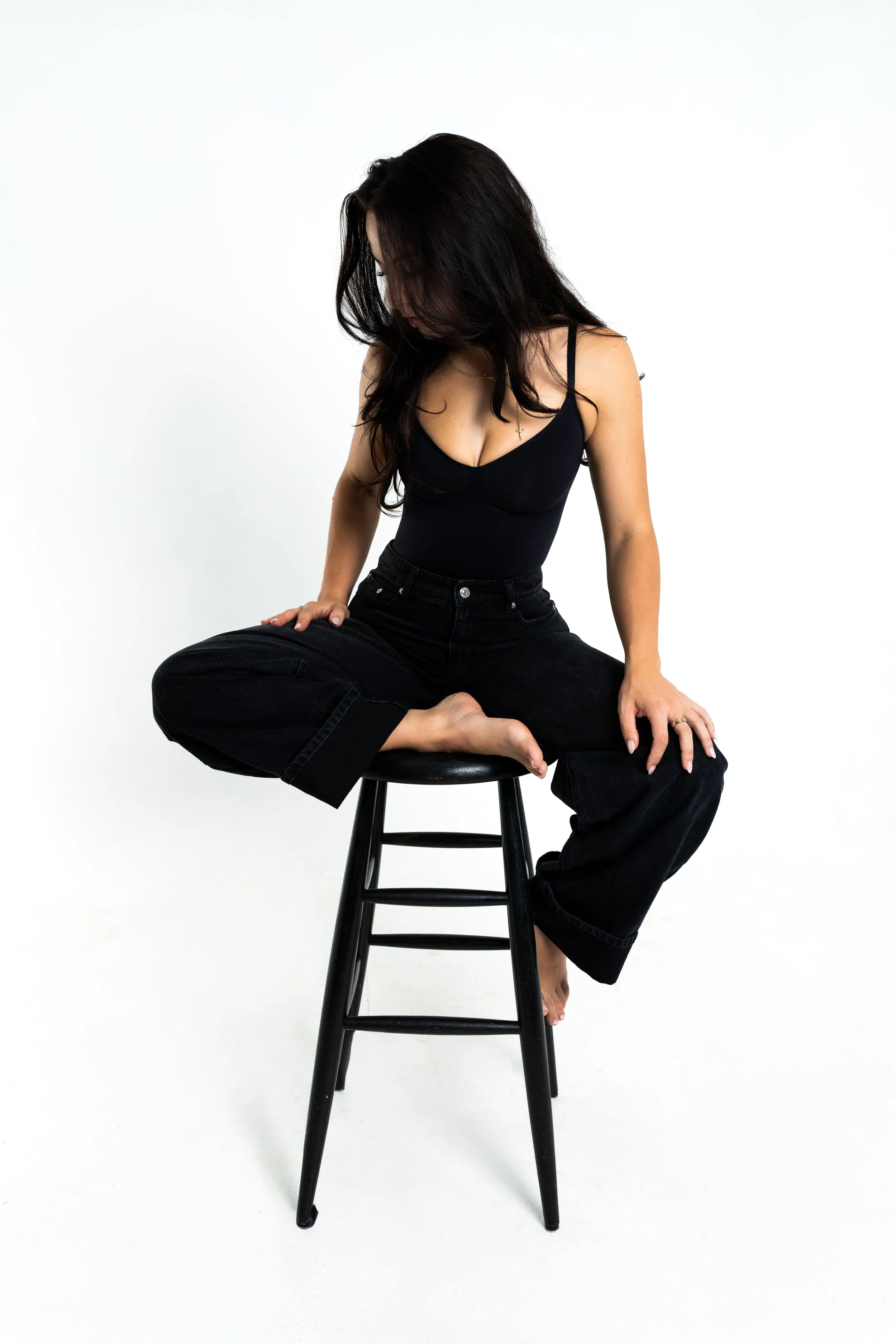 Woman with long dark hair wearing black top and black pants sitting cross-legged on a stool against a white background, looking down.