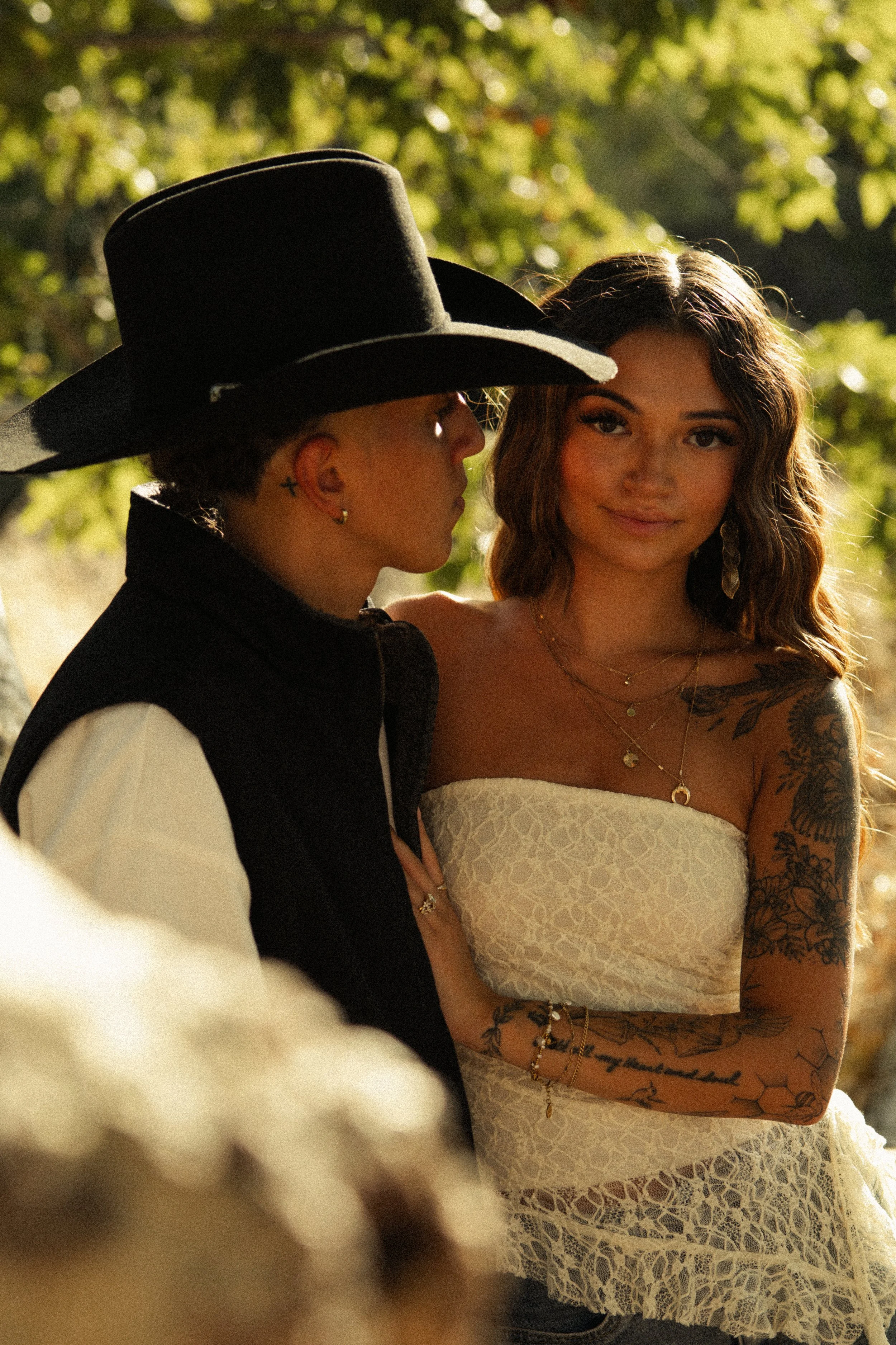A woman with long, wavy hair and tattoos on her arms, wearing a white lace dress with jewelry, standing outdoors with sunlight filtering through trees. Next to her, a person with short hair, earrings, a cross earring, and a large black hat is partial