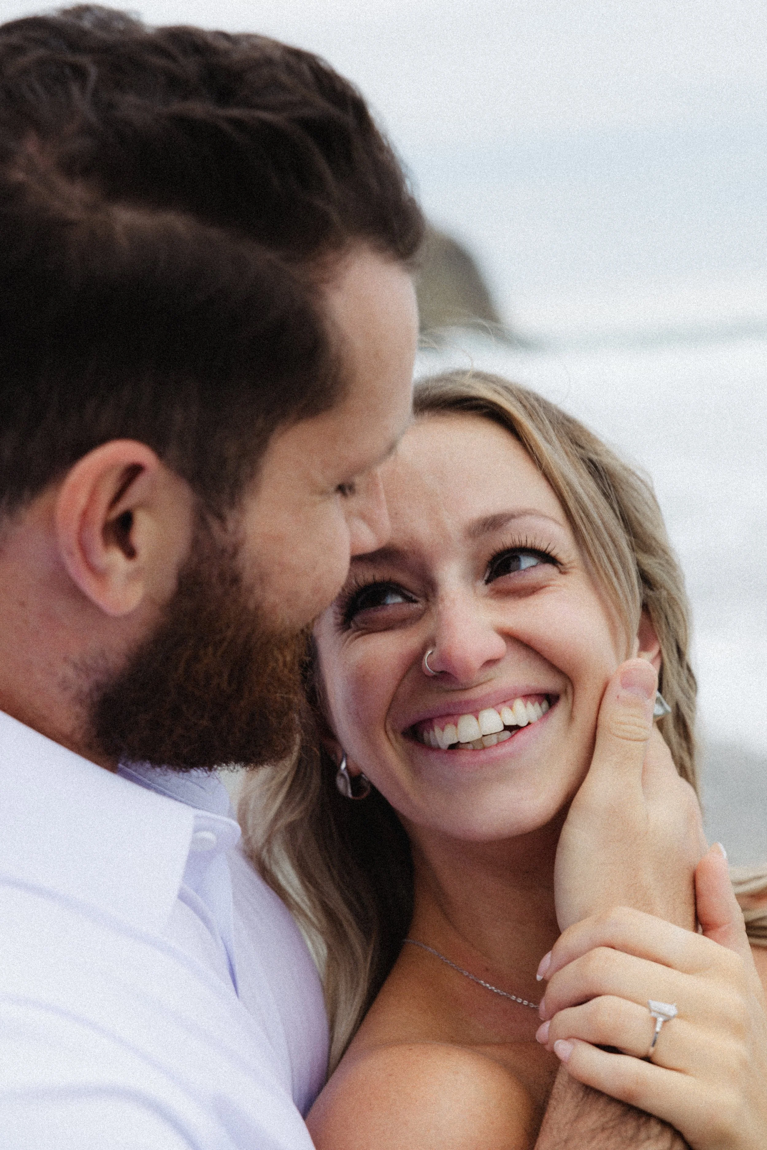 A close-up photo of a man and woman smiling and cuddling closely, with the woman looking happy and the man appearing affectionate.