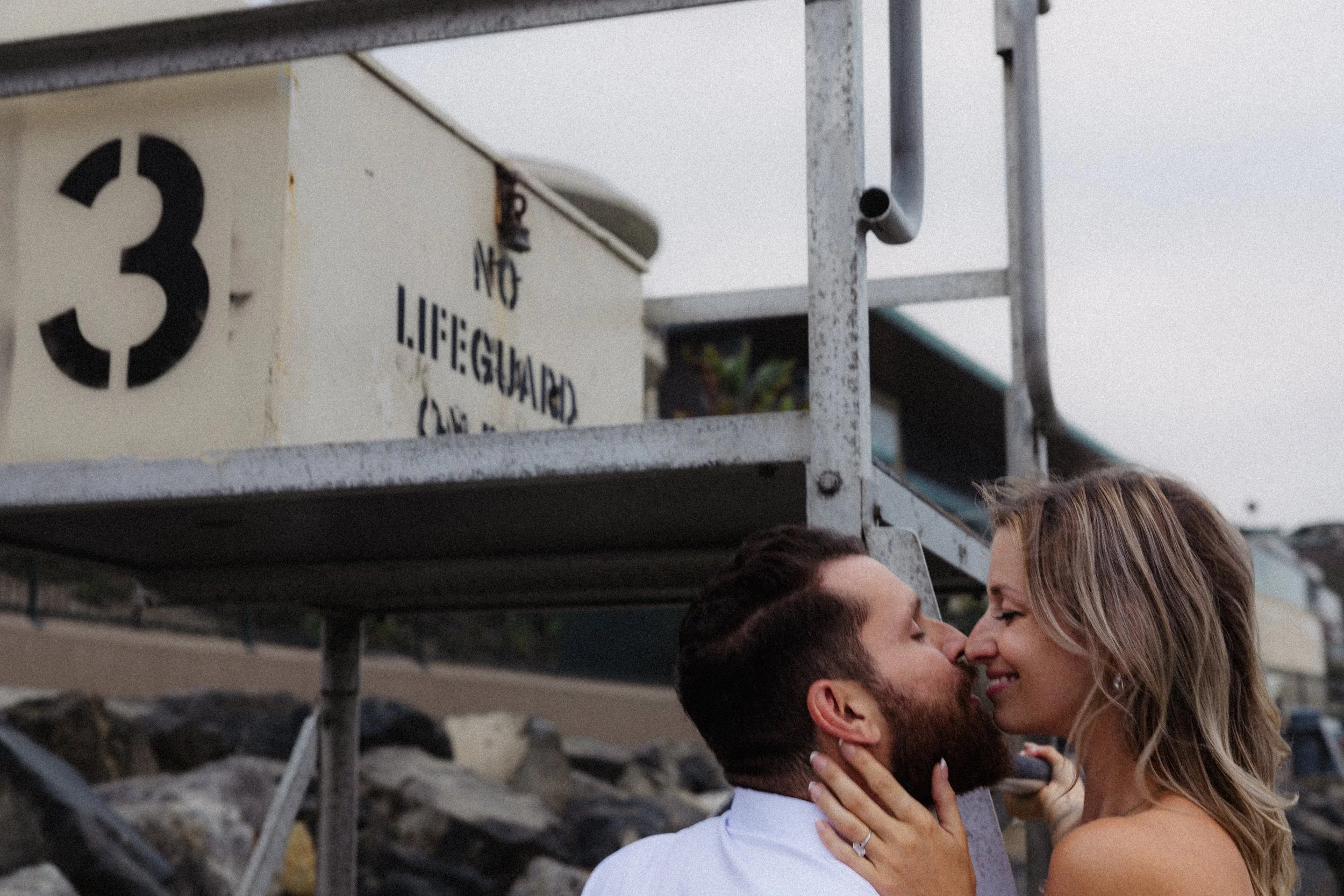 A couple is about to kiss outdoors on a rocky surface, with a metal structure and a sign in the background.