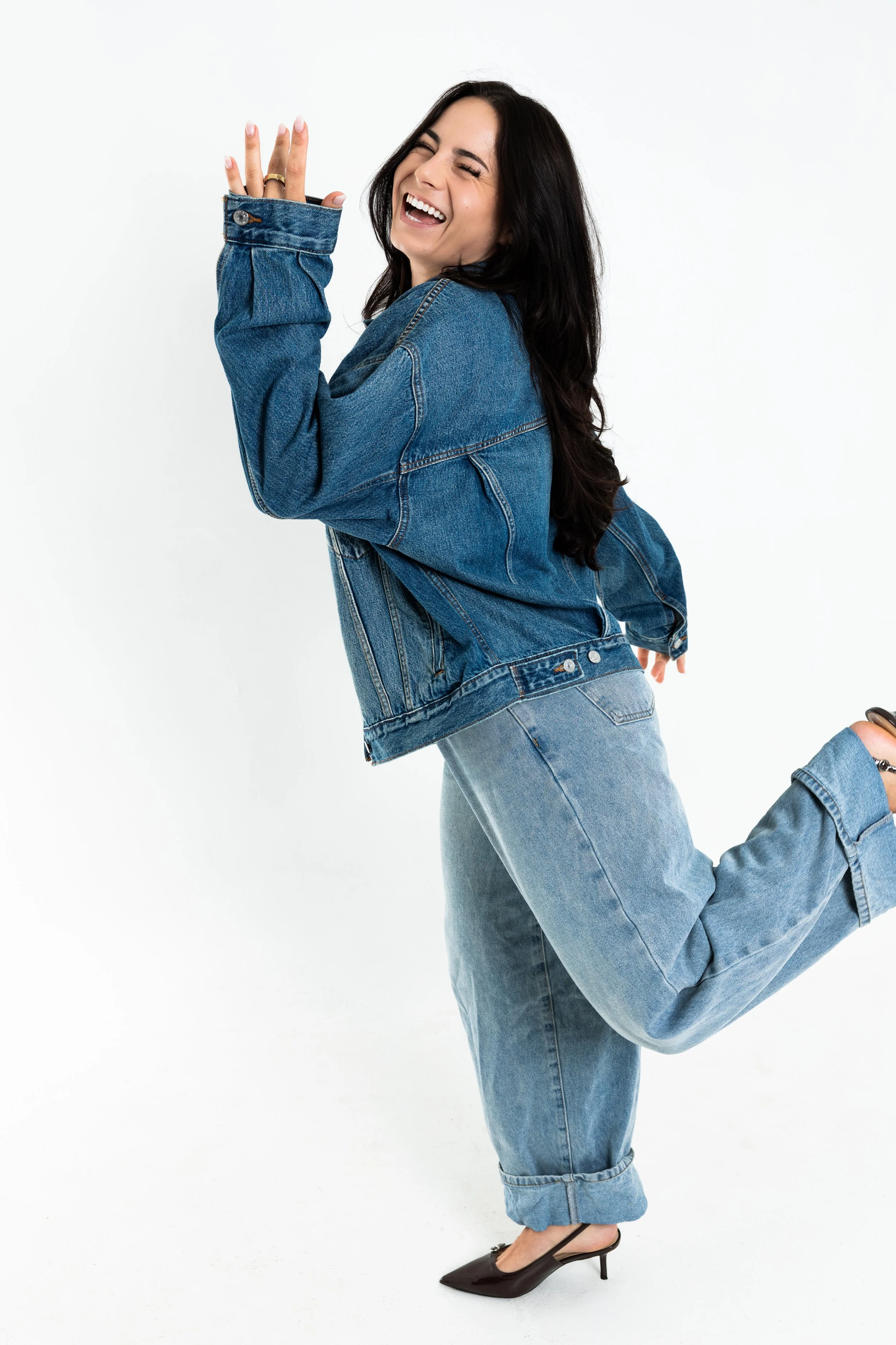 A woman with long dark hair wearing a denim jacket and jeans, black high heels, and smiling while in a playful pose against a plain white background.