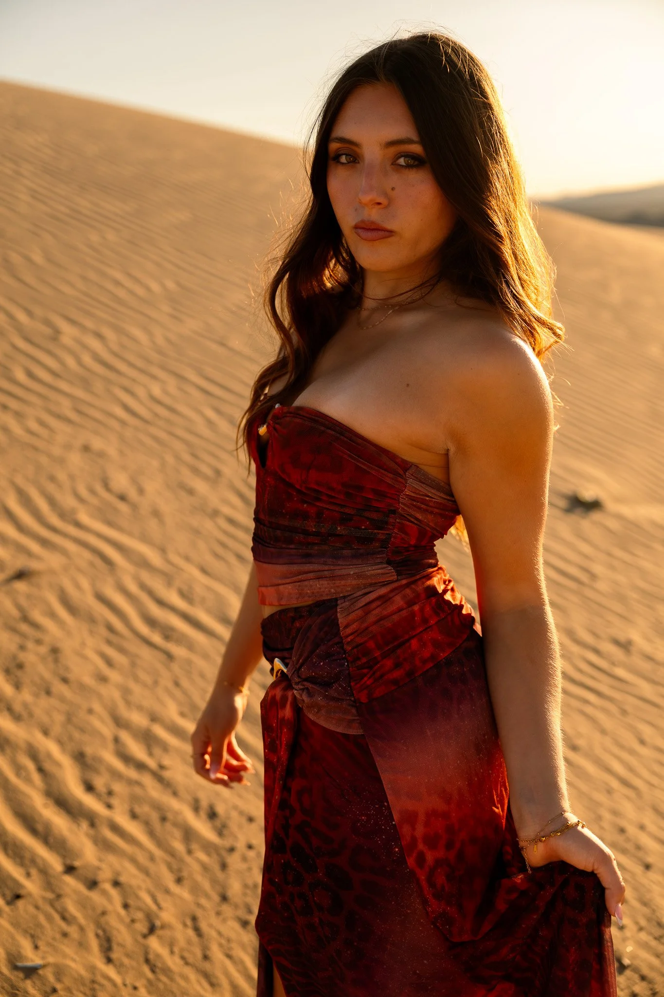 A woman with long brown hair and green eyes wearing a strapless red dress in a desert during sunset.