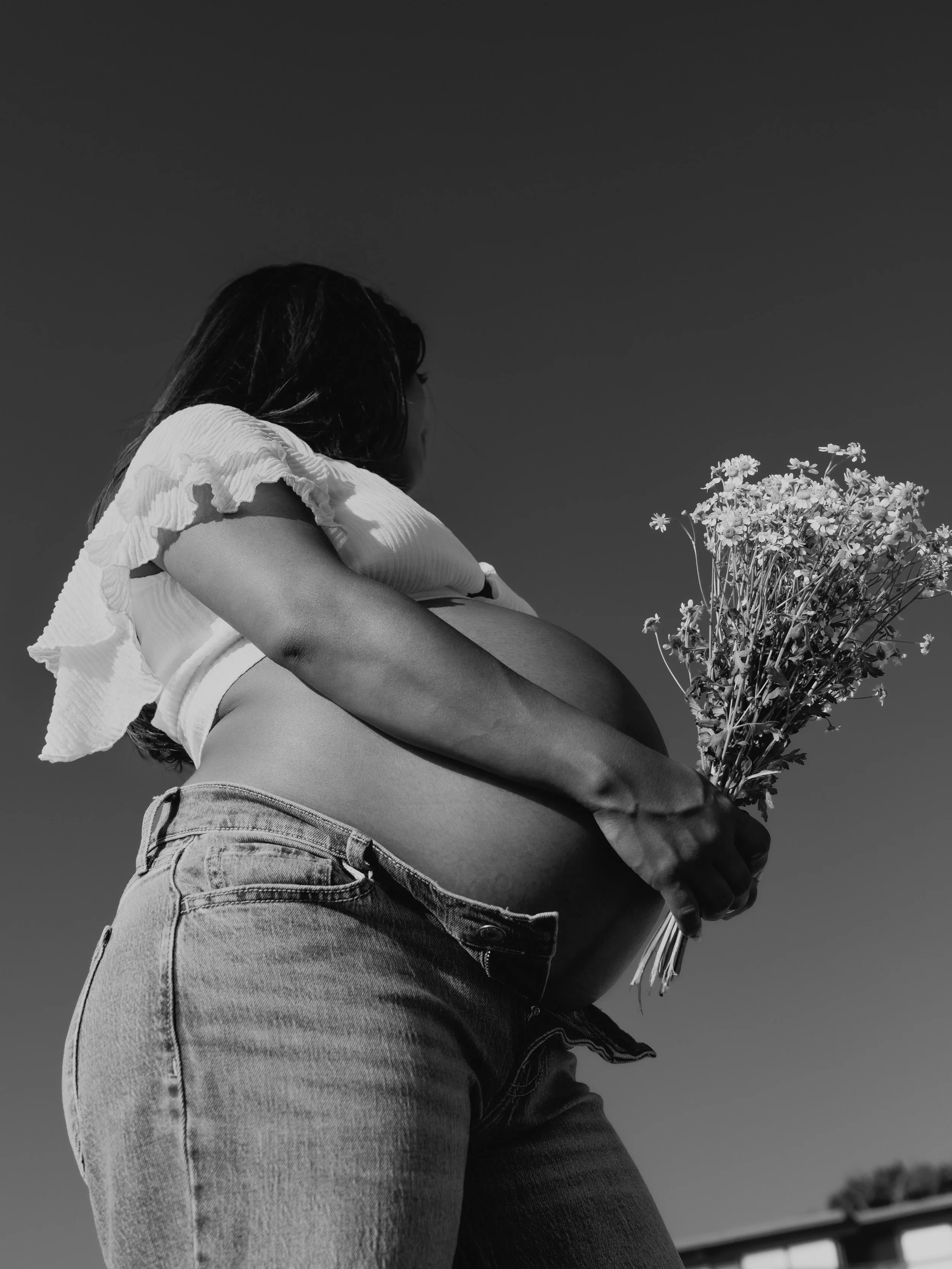 A black and white photo of a person with long hair wearing a short-sleeved shirt and jeans, holding a bouquet of flowers, taken from a low angle under a clear sky.