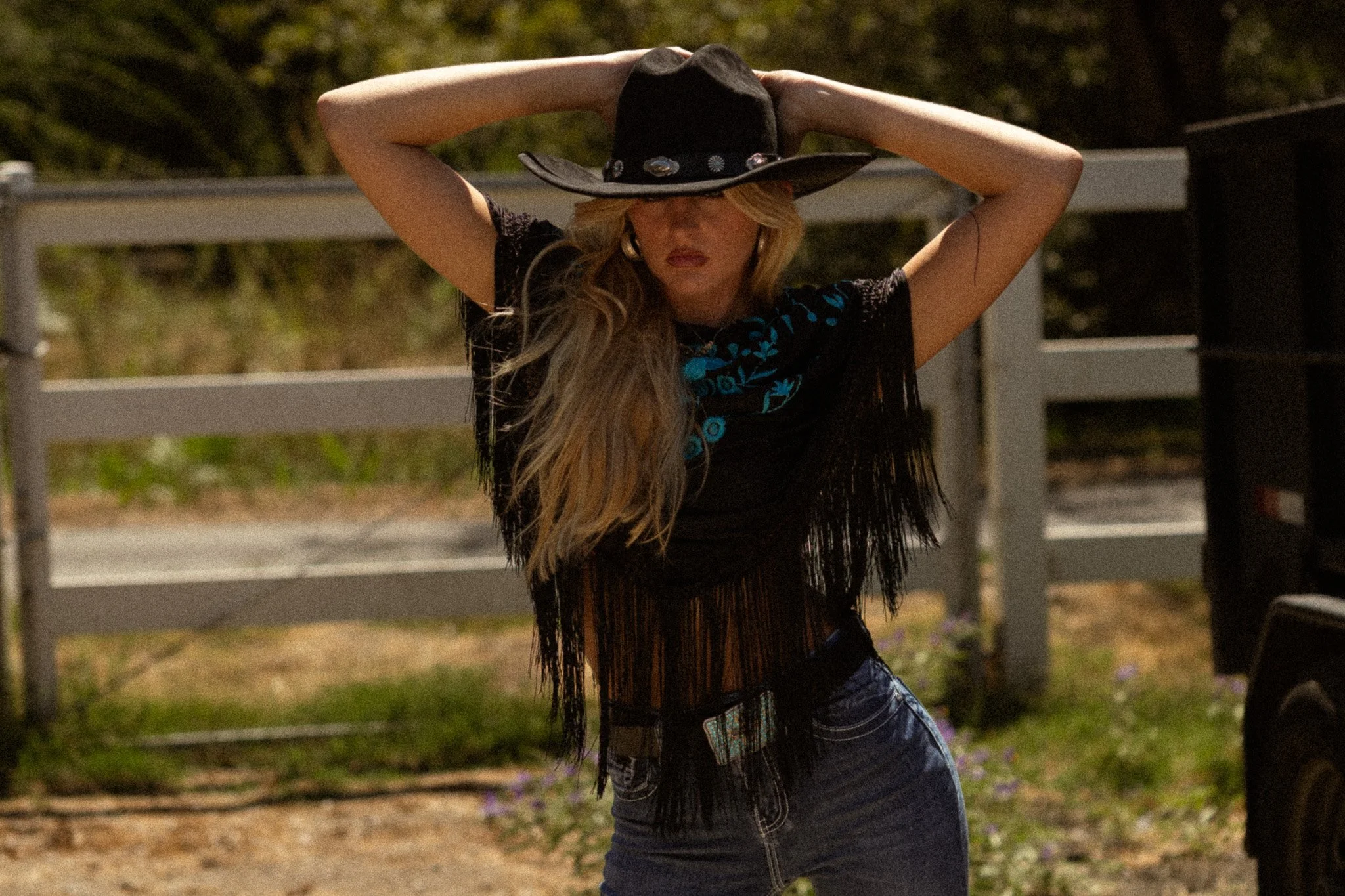 A woman with long blonde hair, wearing a black wide-brimmed hat decorated with conchos, a black top with blue embroidery, and blue jeans, stands outdoors with her arms raised above her head against a background of trees and a white fence.