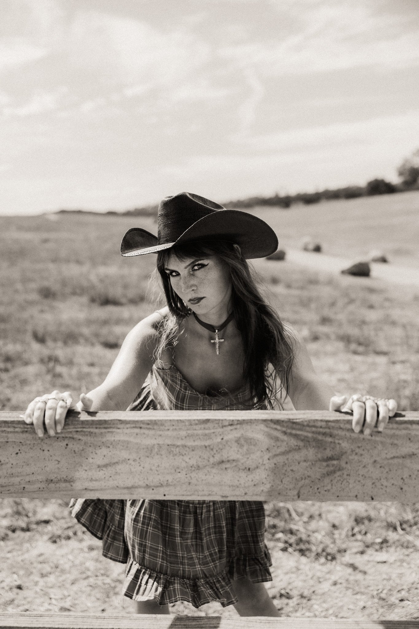 A woman wearing a large cowboy hat and plaid outfit, standing behind a wooden fence in a desert landscape with a cloudy sky.