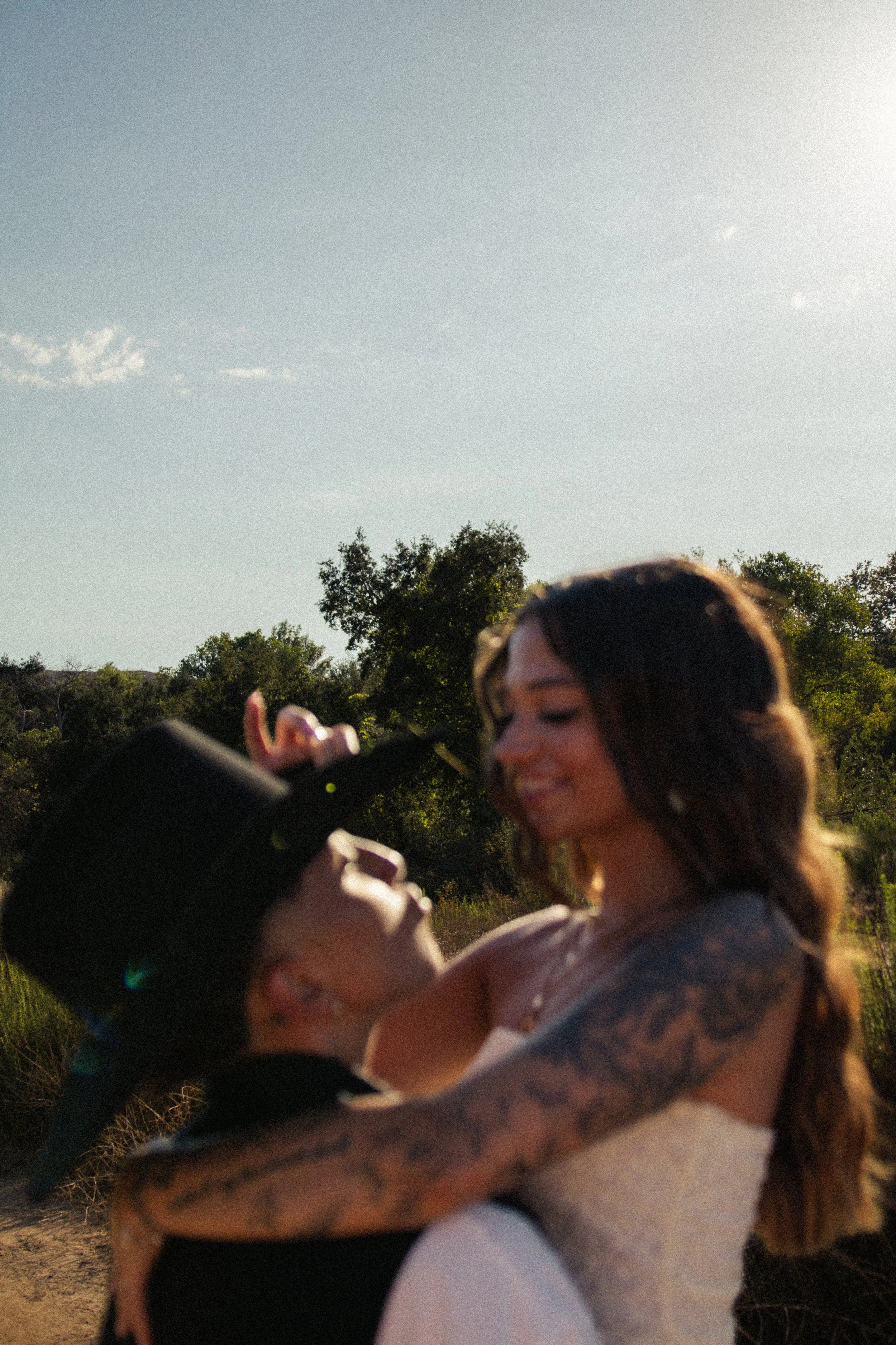 Two women embrace outdoors with trees and a clear sky in the background, one woman has tattoos and is smiling, the other woman has dark hair and is holding a phone.