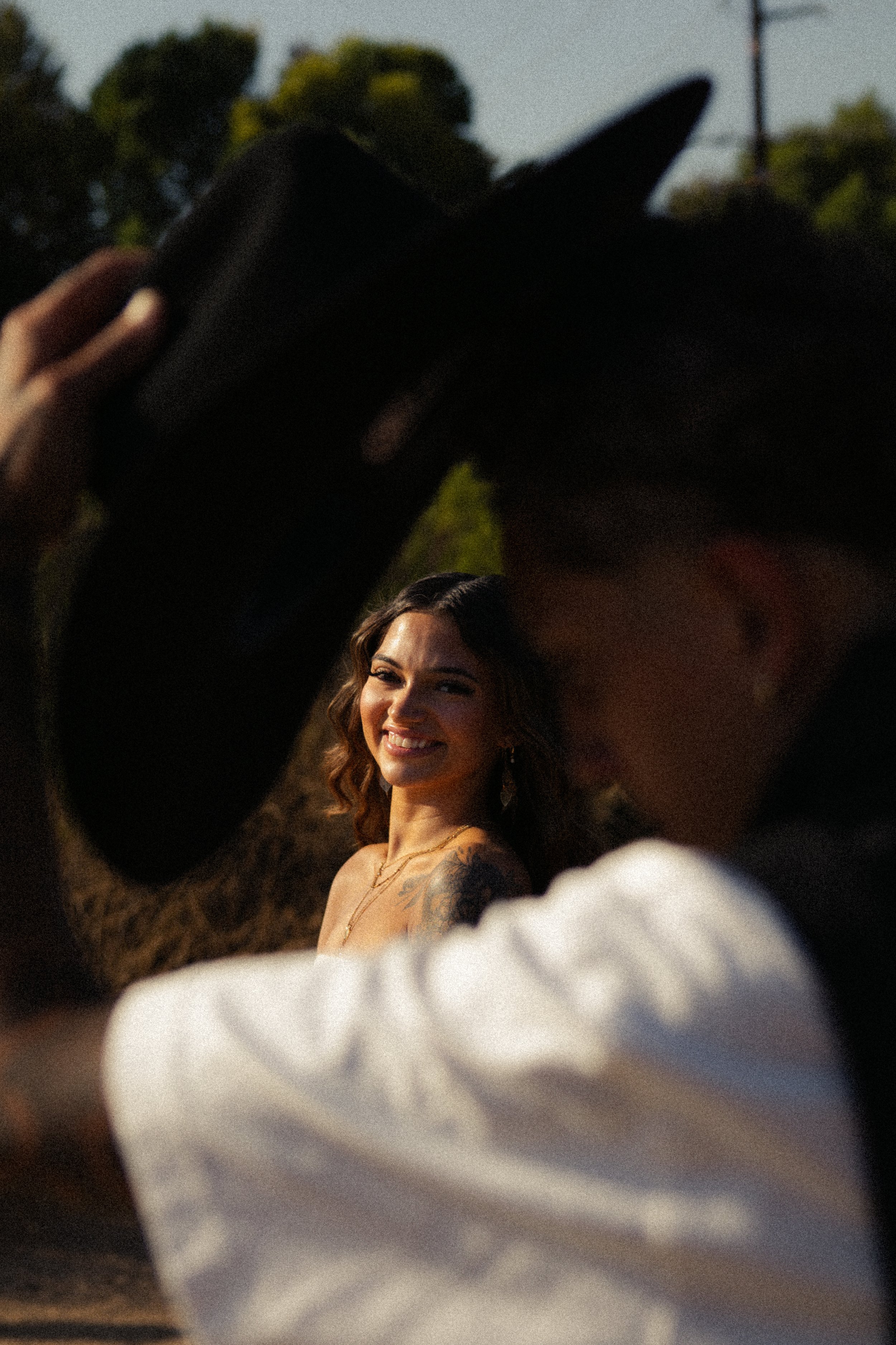 A woman with long wavy hair and a tattoo on her left shoulder poses outdoors, smiling, while two people in the foreground hold a black hat above her head during sunset or golden hour.