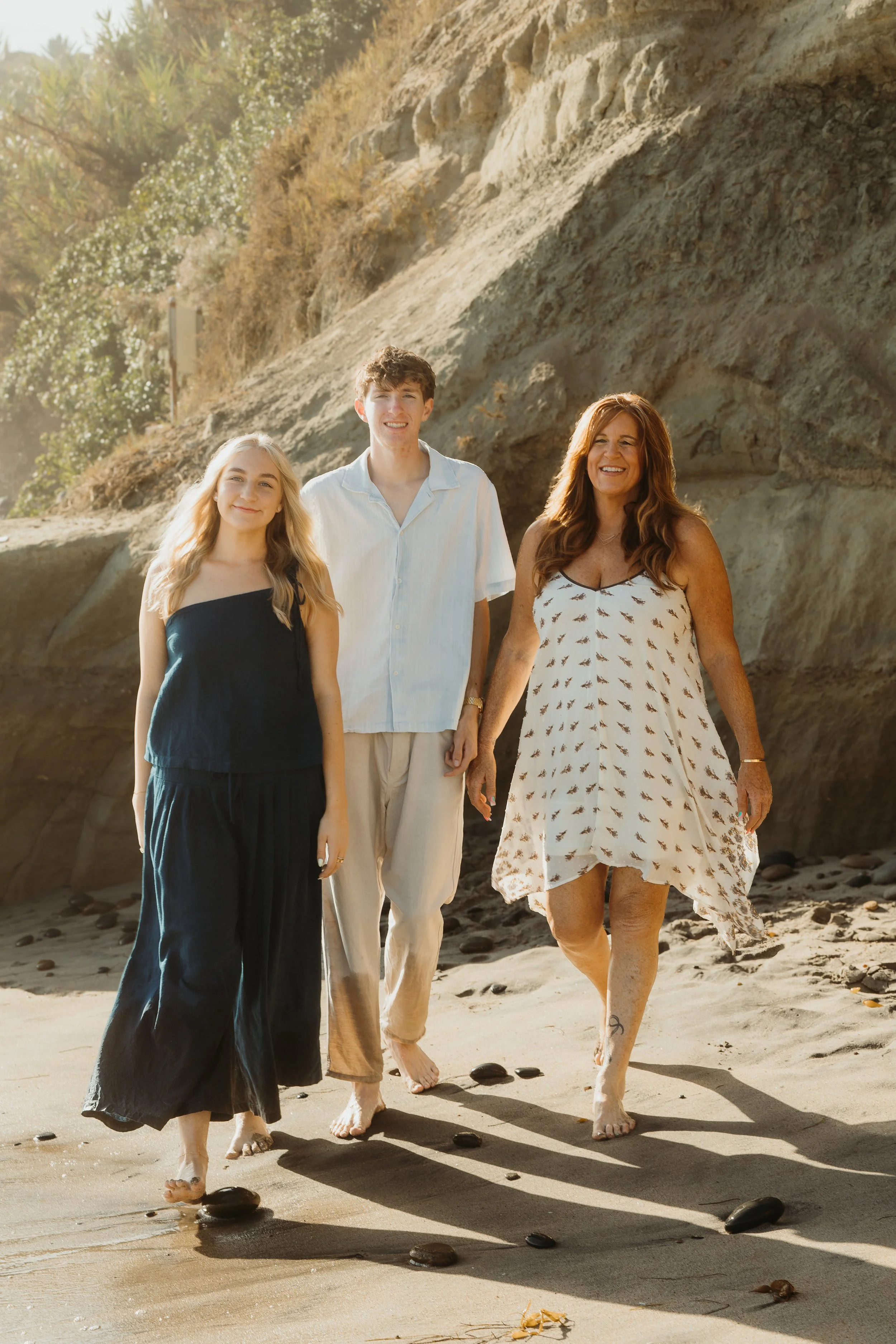 Three women and one man walking on a beach with rocks and cliffs in the background. The women are smiling, and the group appears to enjoy a sunny day.