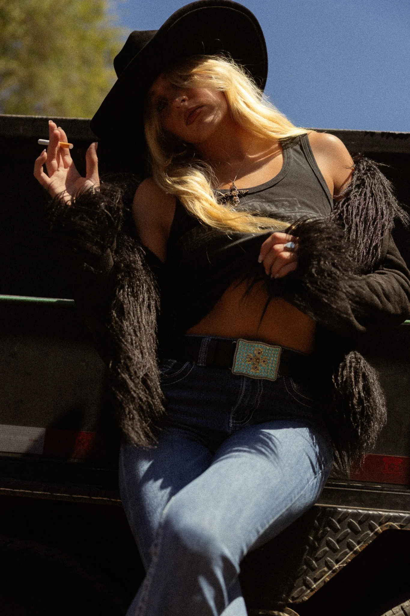 A woman with blonde hair wearing a black hat, black tank top, and a furry jacket, holding a cigarette, sitting with her legs crossed on a bench outdoors during daylight.
