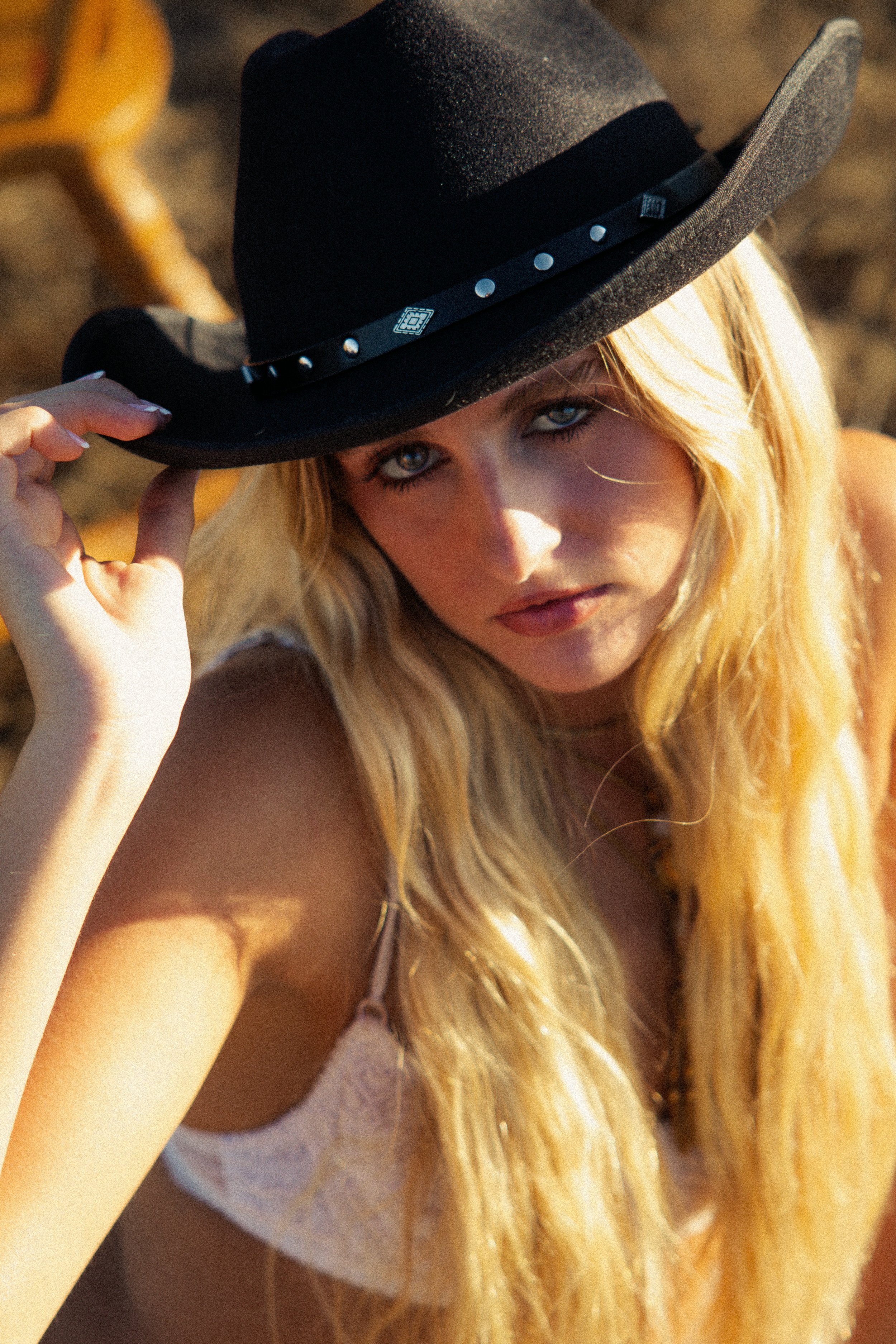A woman with long blonde hair wearing a cowboy hat and a white lace top, holding the brim of her hat.