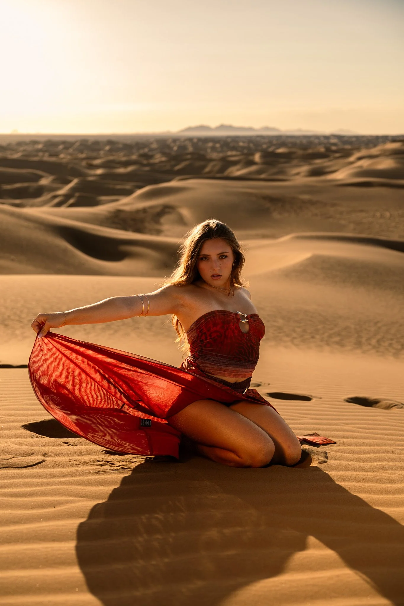A woman in a red dress kneeling on sand dunes in a desert during sunset, holding the edge of her dress with one arm extended.