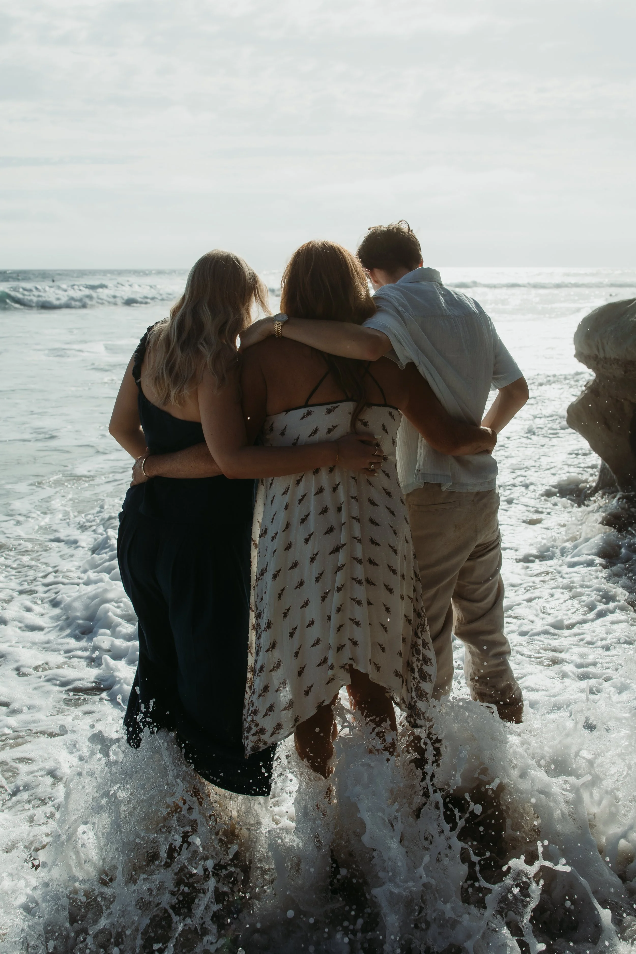 Three people walking in the ocean with waves splashing around their legs, embracing each other, with the sea and sky in the background.