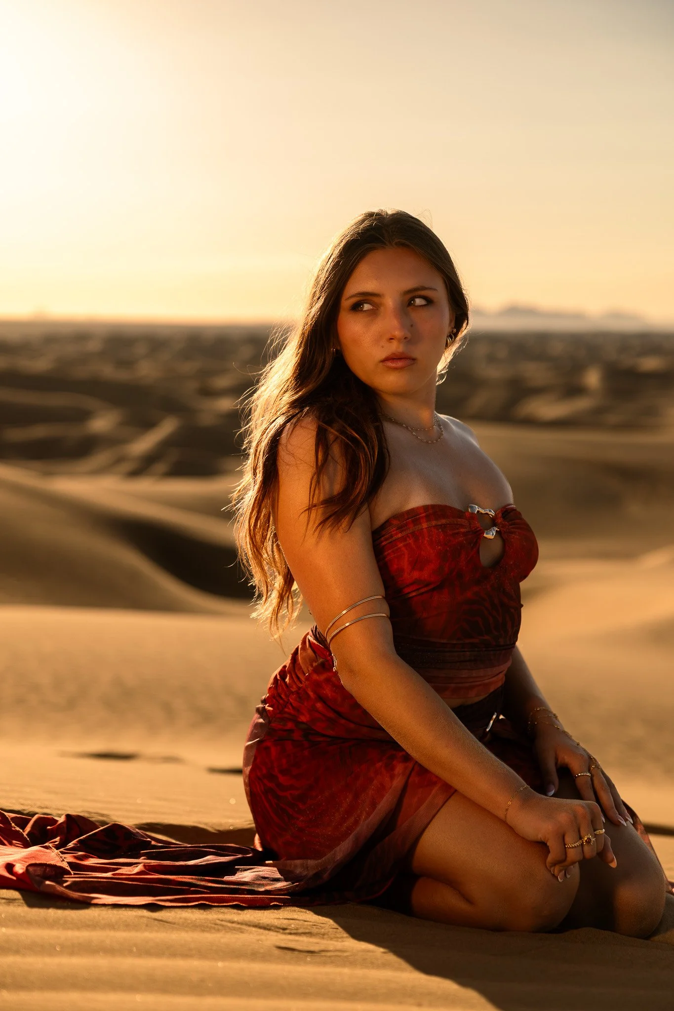 A woman with long brown hair and light skin sitting on the desert sand during sunset, wearing a red strapless dress and jewelry.