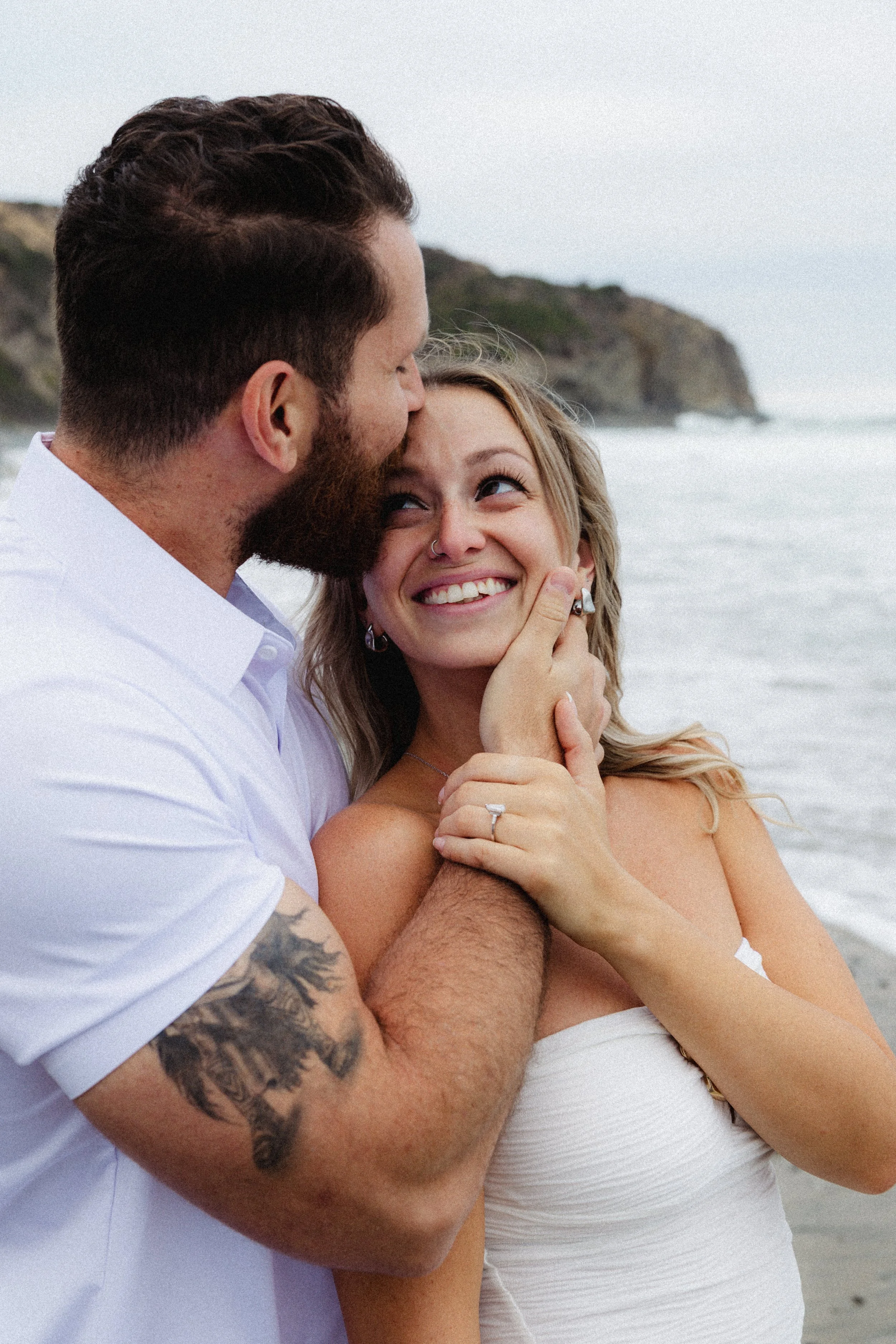 A man with a beard and tattoos on his arm kissing a smiling woman on a beach, with cliffs in the background.
