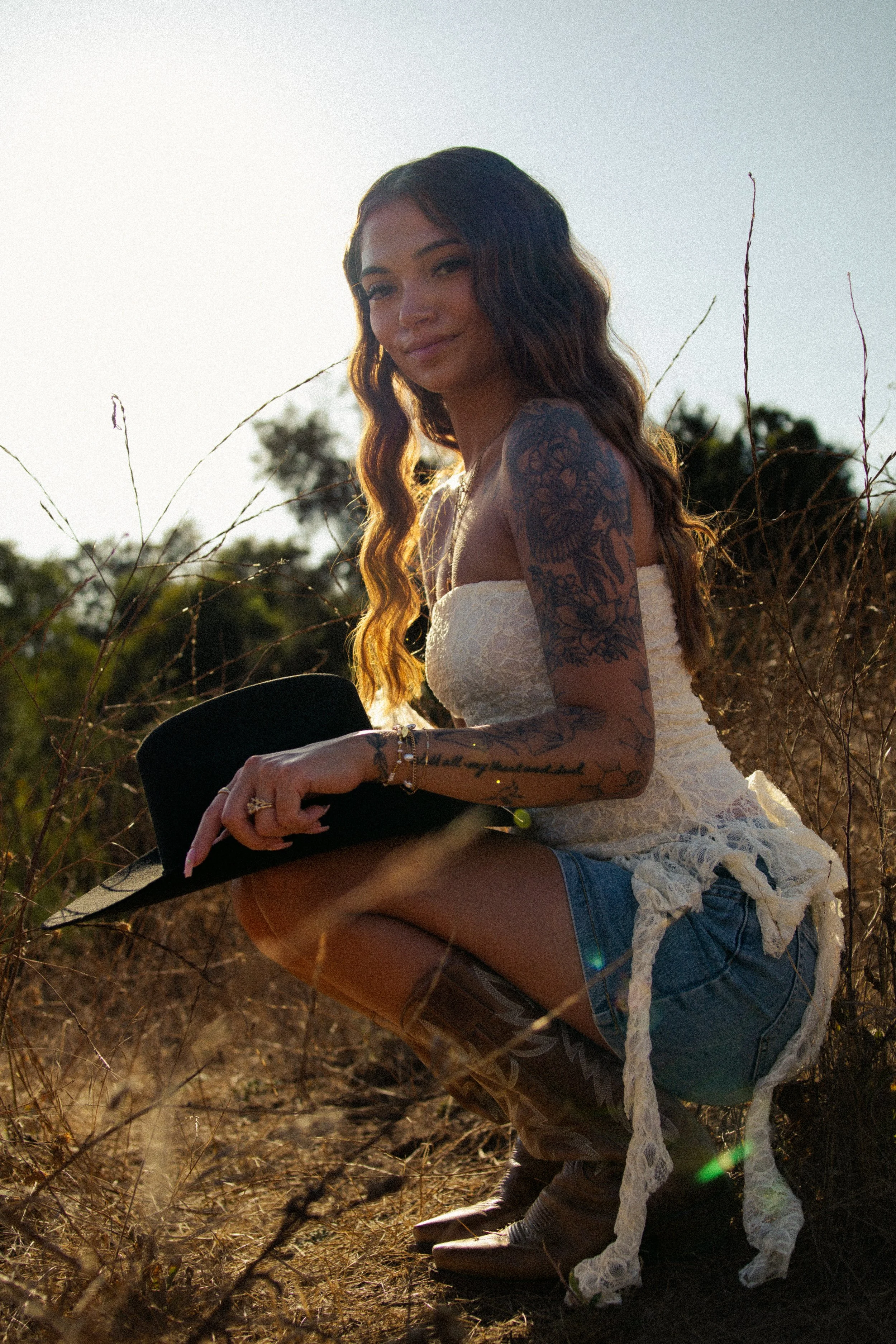 A woman with long wavy hair and tattoos on her arm is outdoors in a field during sunset, wearing a white top, shorts, and cowboy boots. She is squatting and holding a black hat, with dry grass and trees in the background.