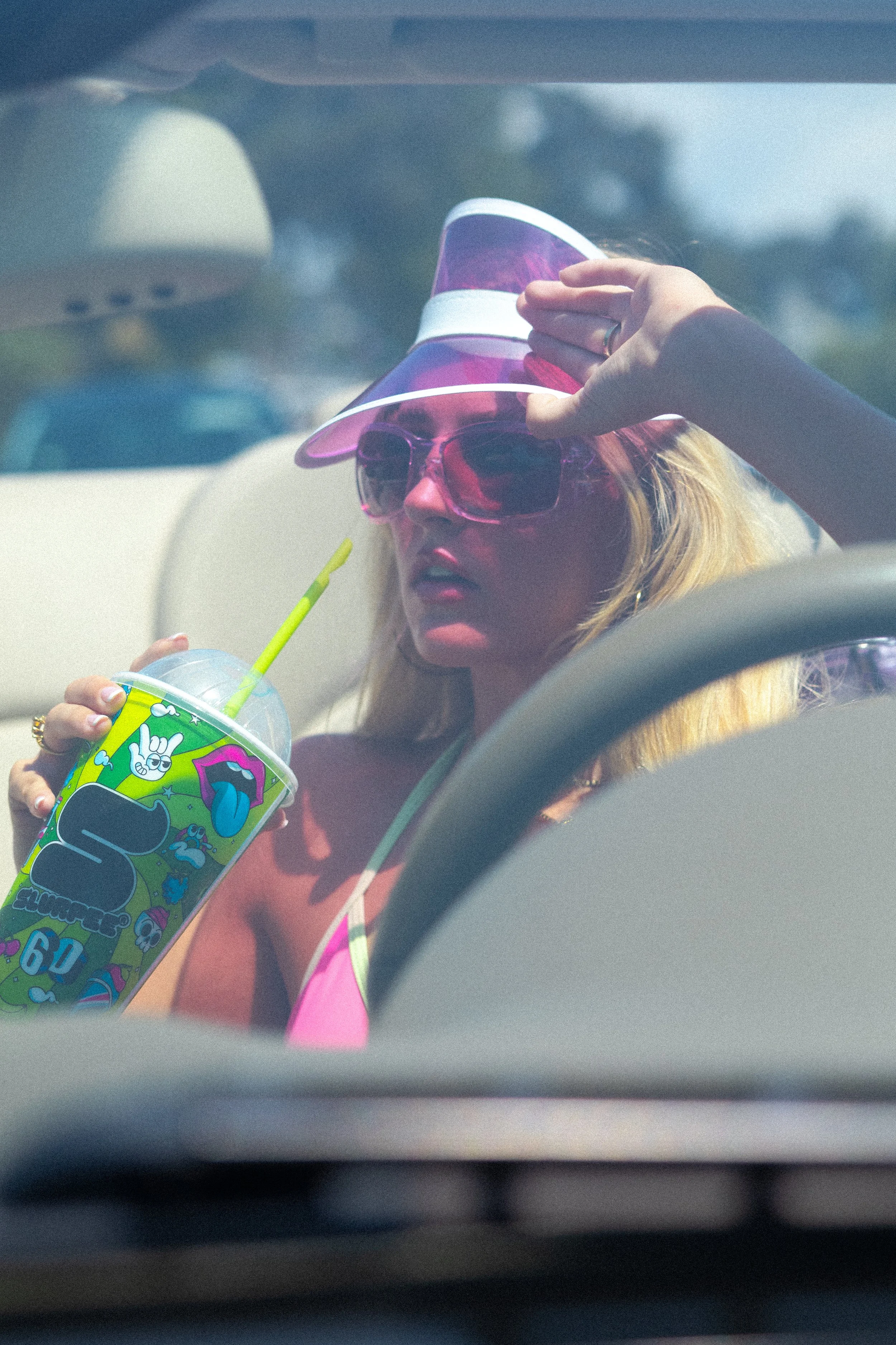 A woman wearing pink sunglasses, a purple visor, and a pink top is sitting in a car, holding a beverage with a green straw, while adjusting her visor with her right hand.