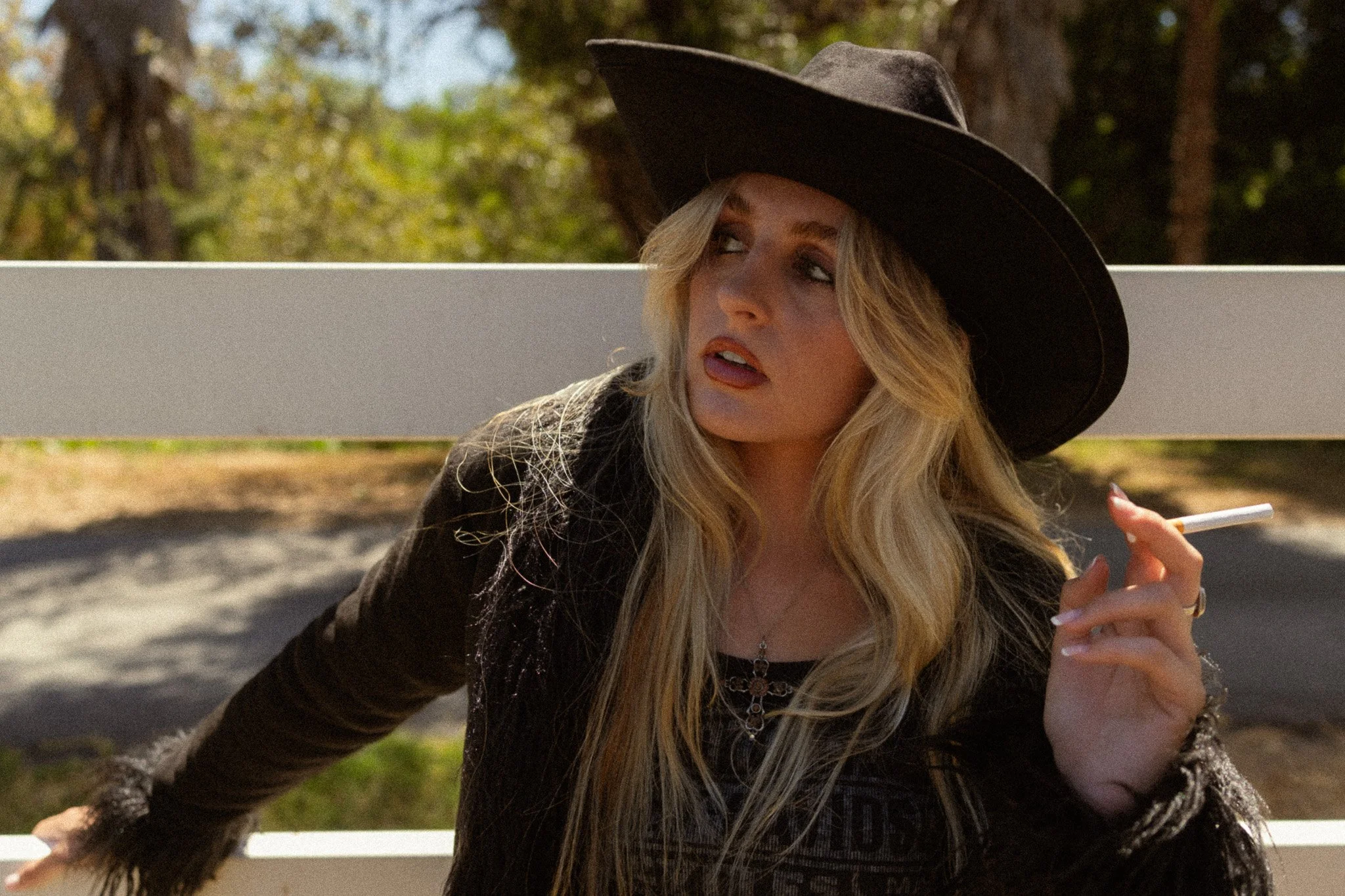 A woman with long blonde hair wearing a large black hat and a black jacket, holding a cigarette, sitting outdoors near a white railing, with trees and green foliage in the background.
