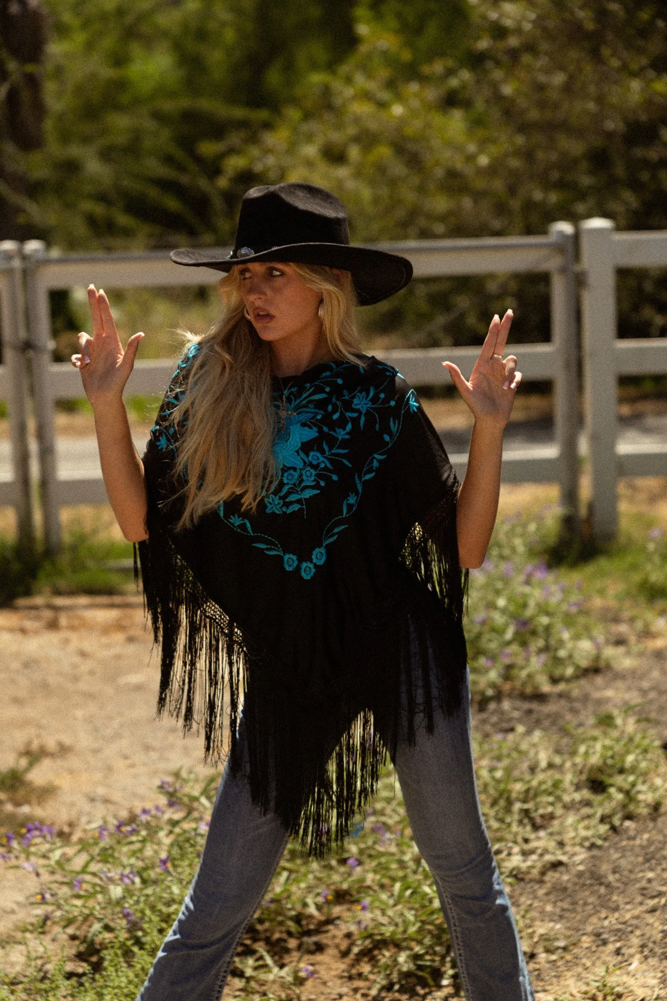 A woman wearing a black wide-brimmed hat, black fringe shawl, and blue floral embroidery top, standing outdoors with a wooden fence and greenery in the background, striking a pose with hands raised.
