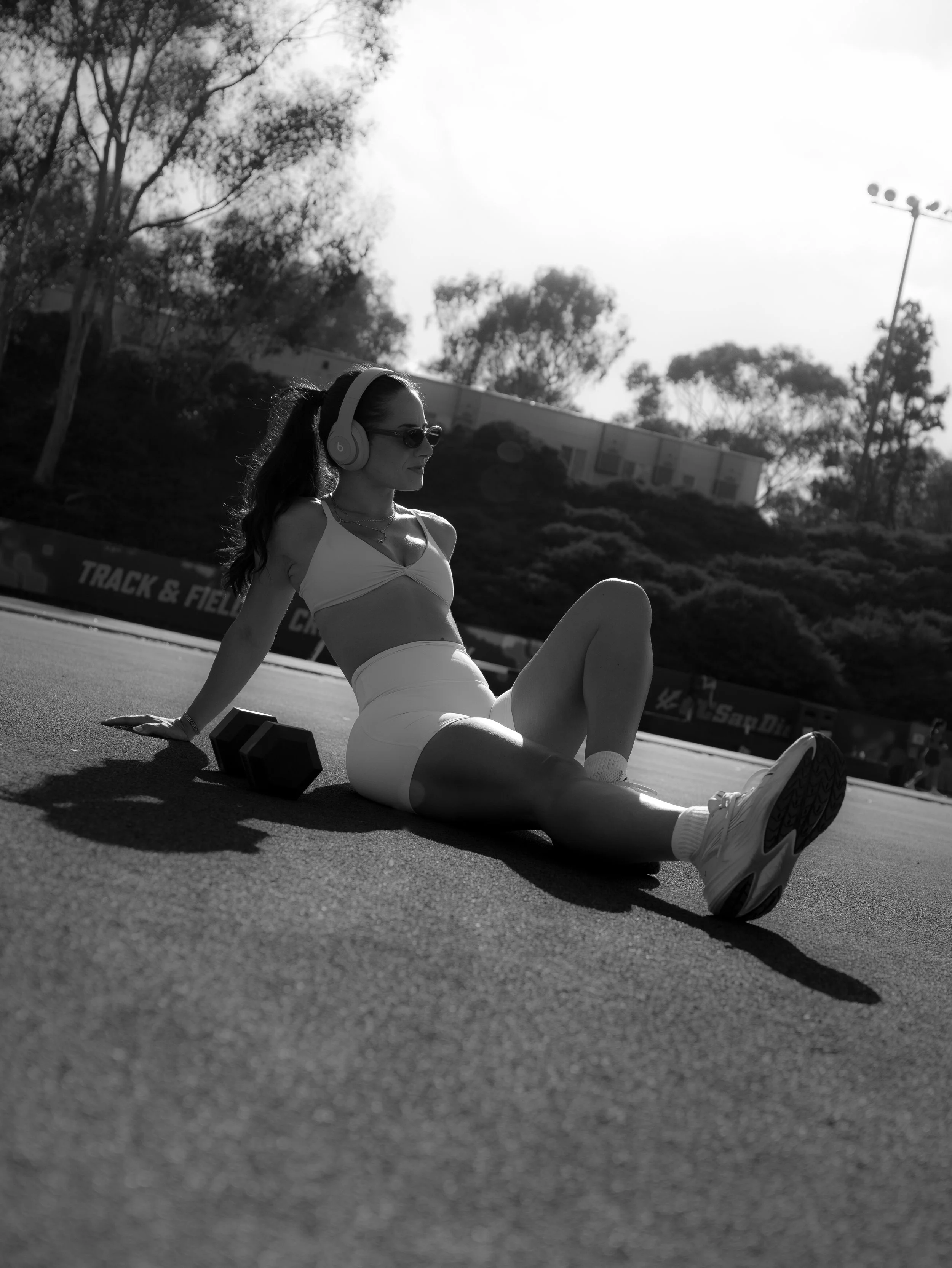 A woman wearing sunglasses, headphones, a sports bra, and shorts sitting on an outdoor track or field with one leg extended and the other bent, with dumbbells nearby and trees in the background.