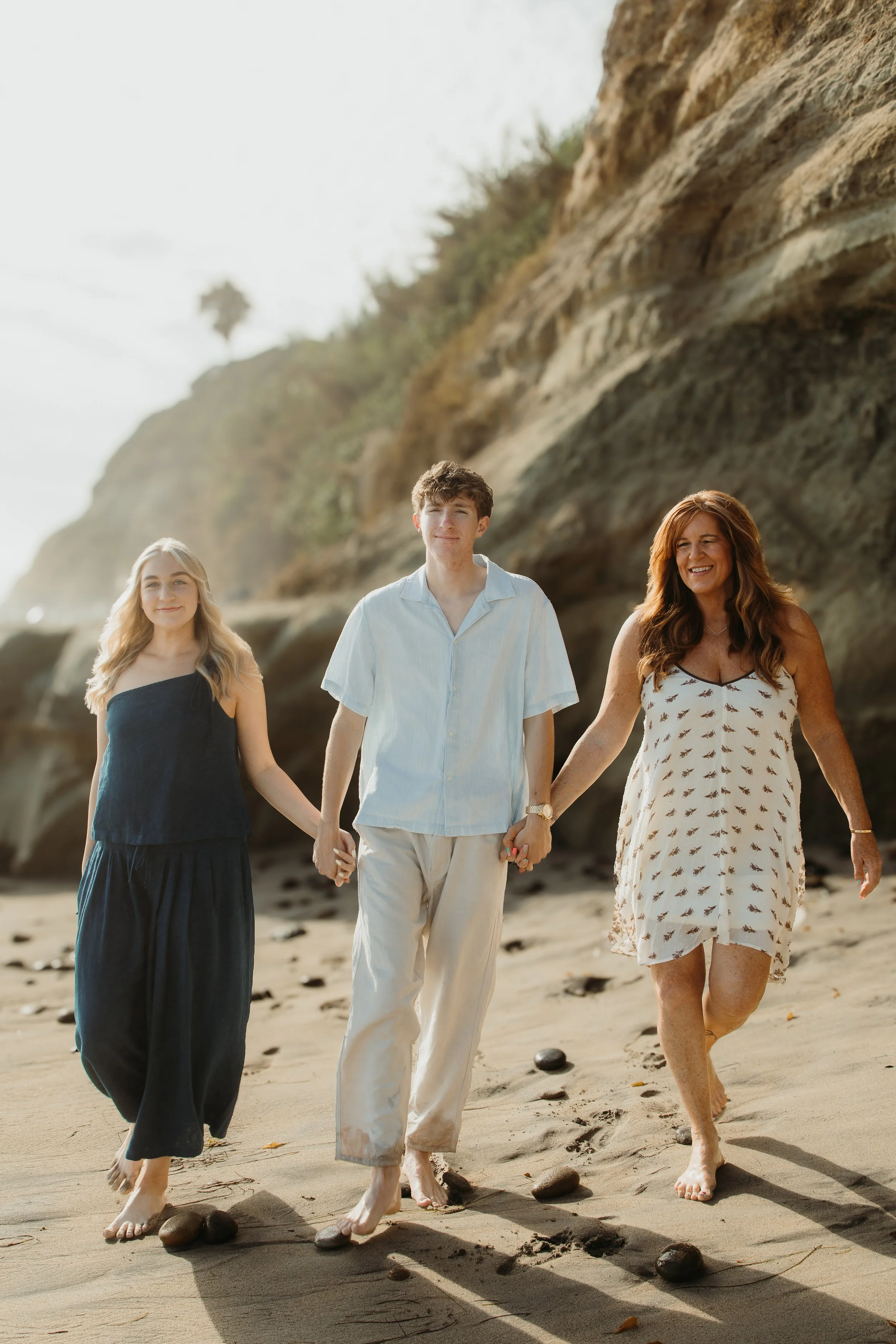 A family of three walking hand in hand along the beach, with rocky cliffs in the background on a sunny day.