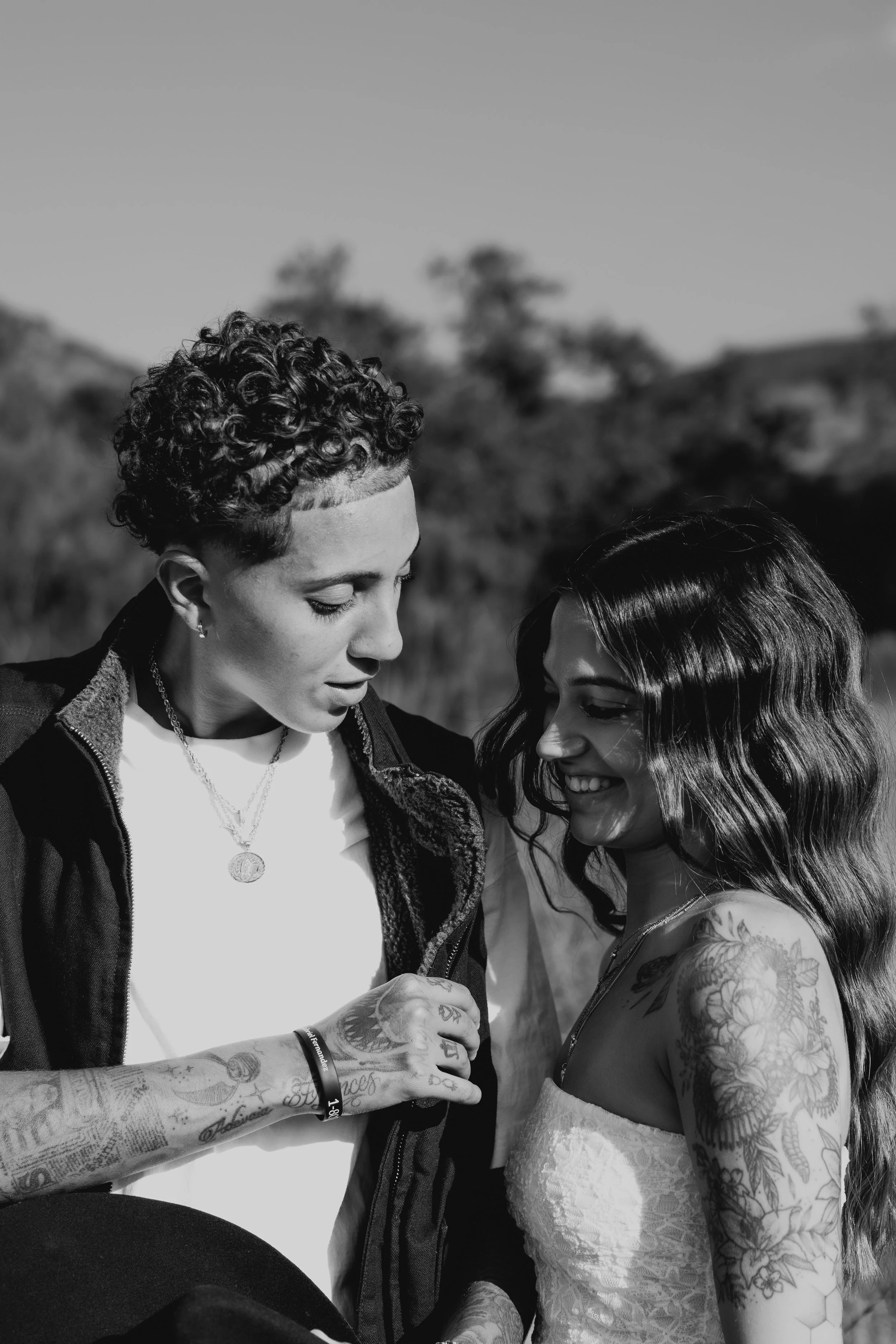 Two women standing close together outdoors, one with curly short hair and tattoos on her arm, the other with long dark wavy hair, both smiling and looking at each other.