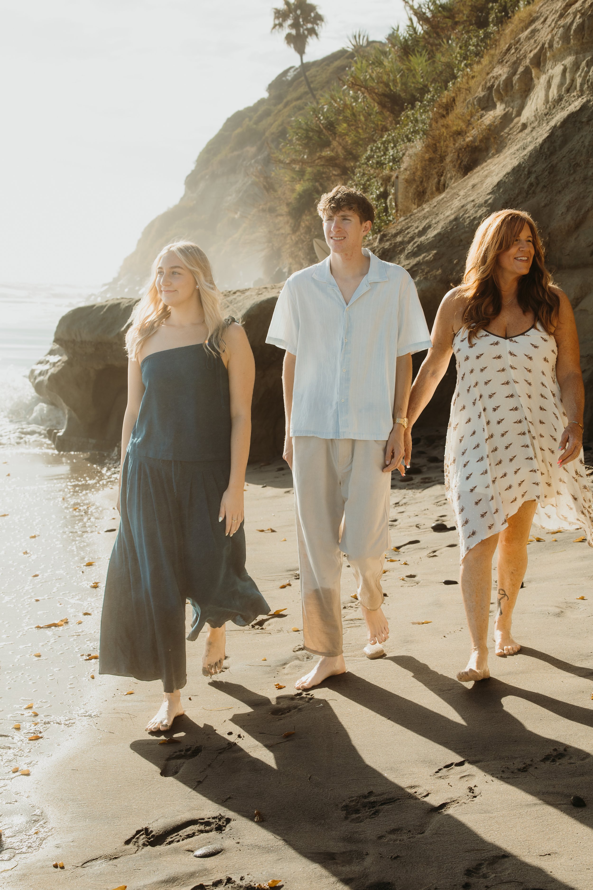 Four people walking barefoot on a sandy beach, holding hands, with cliffs and palm trees in the background on a sunny day.