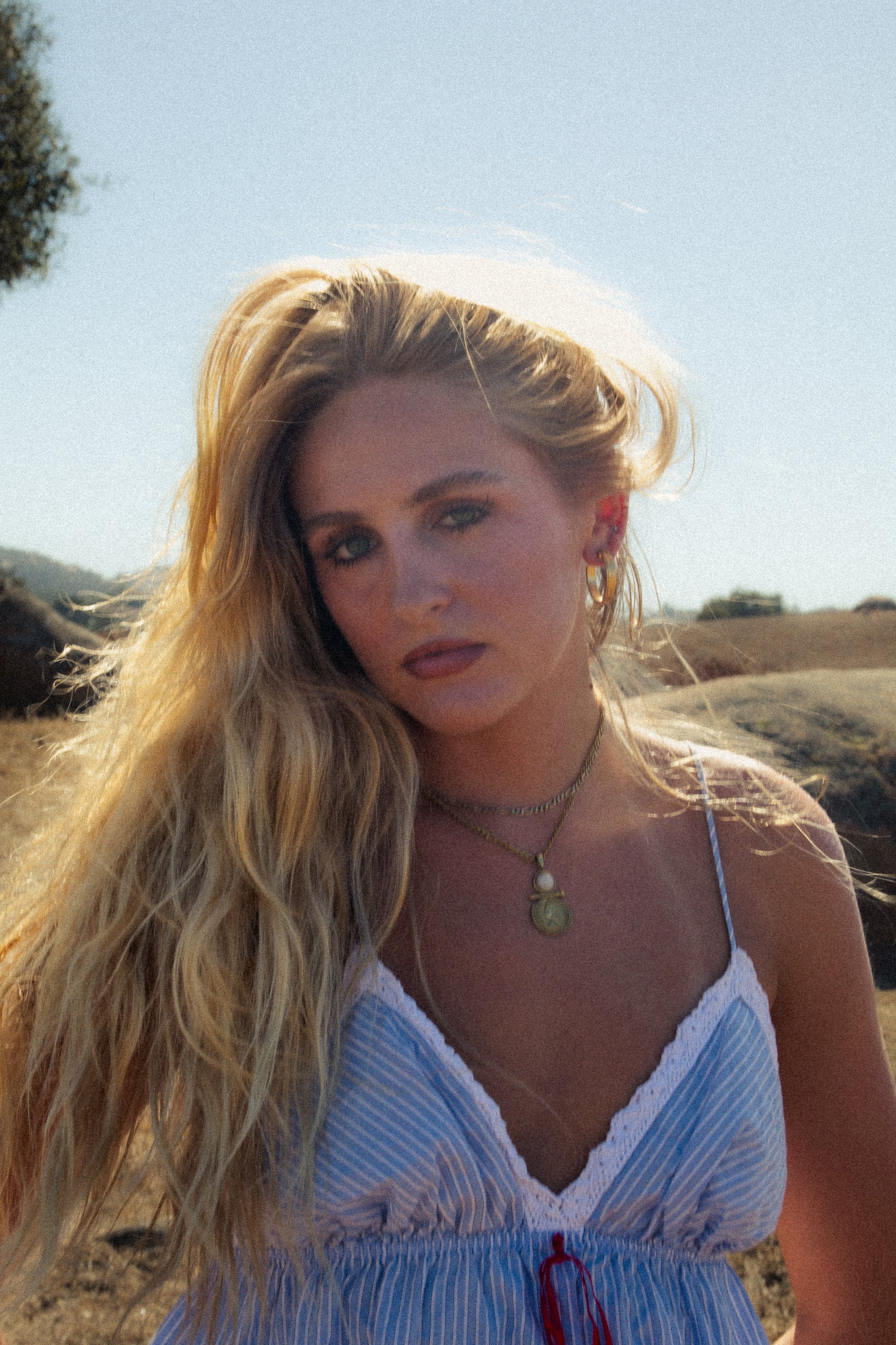 Close-up of a woman with long blonde hair, wearing jewelry, standing outdoors on a sunny day with a blue sky and landscape in the background.
