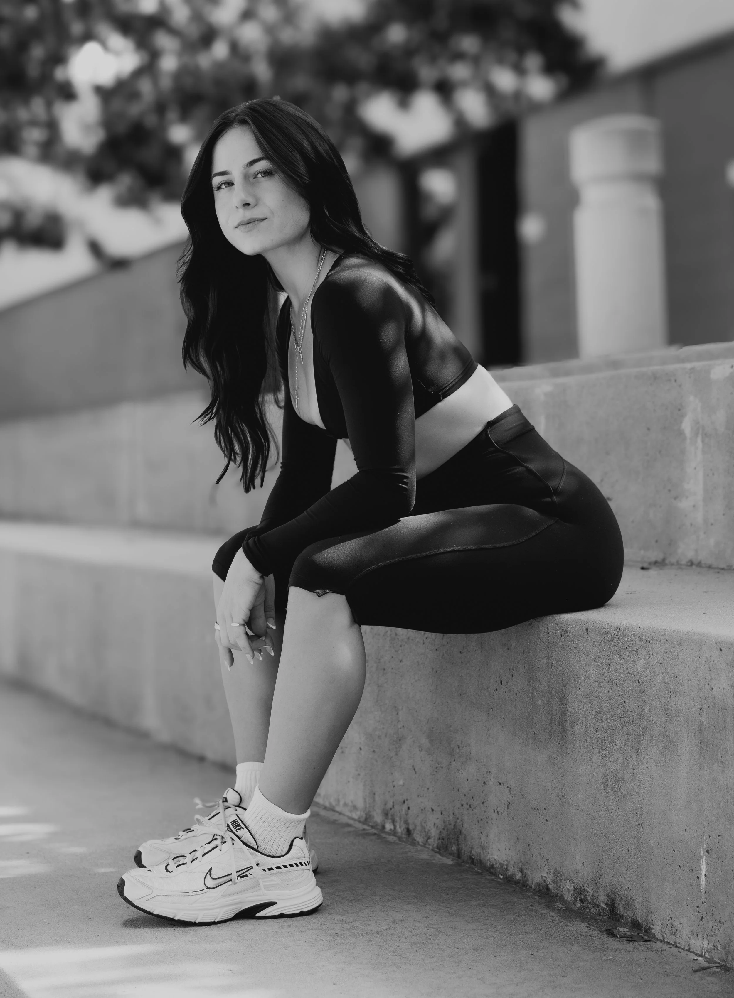 A black-and-white photo of a young woman sitting on a concrete ledge outdoors. She is wearing athletic clothing, including a crop top, leggings, and sneakers, with socks. She has long dark hair and is looking toward the camera with a slight smile.