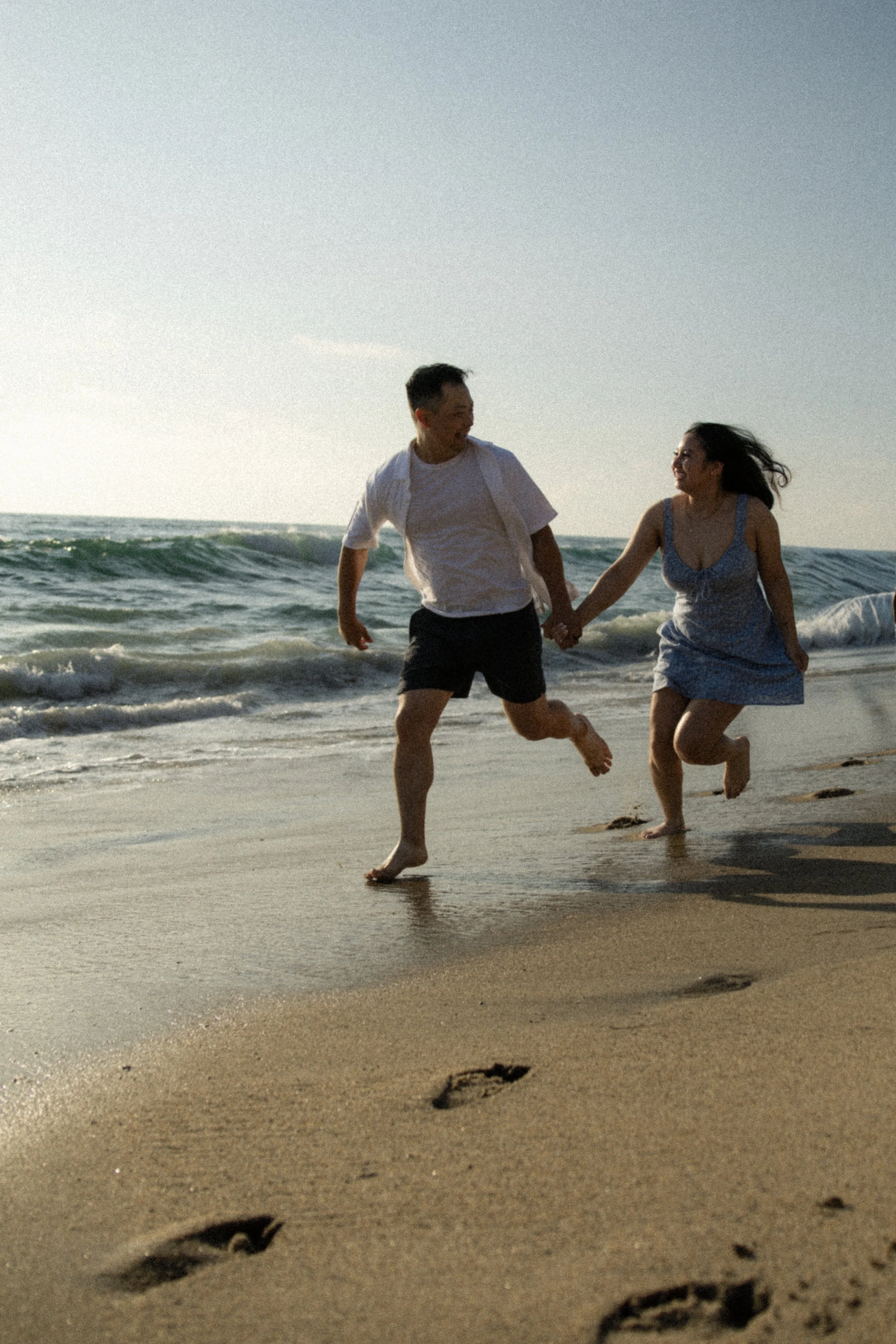 A couple running hand in hand along the shoreline of the beach with footprints in the foreground and ocean waves in the background during sunset.
