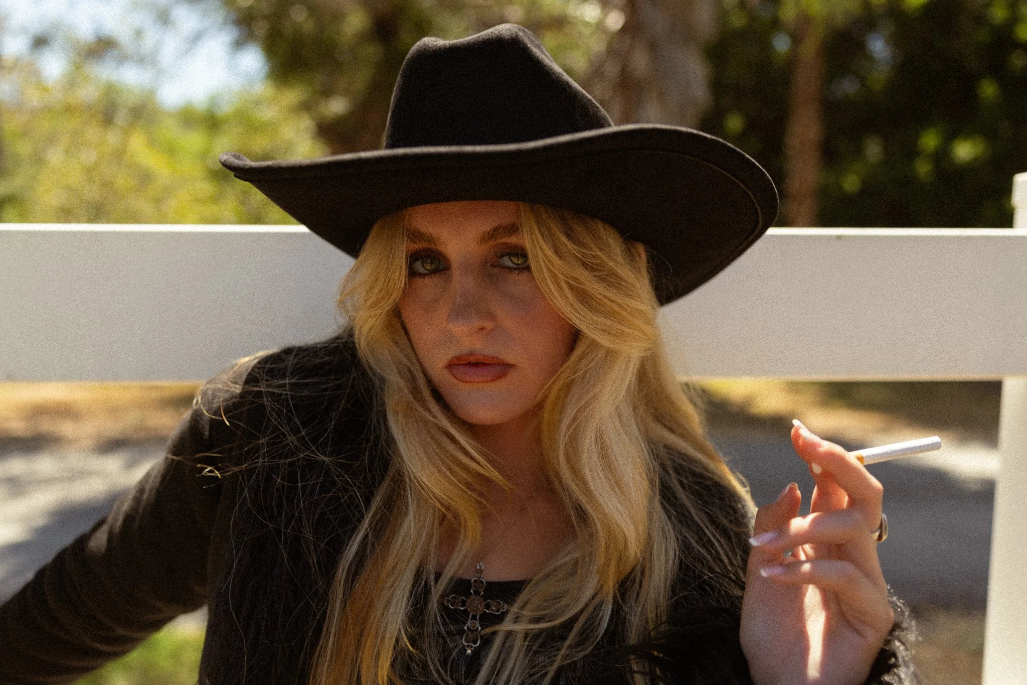 A woman with long blonde hair wearing a black wide-brimmed hat and black clothing, holding a cigarette in her hand, outdoors with green trees in the background.