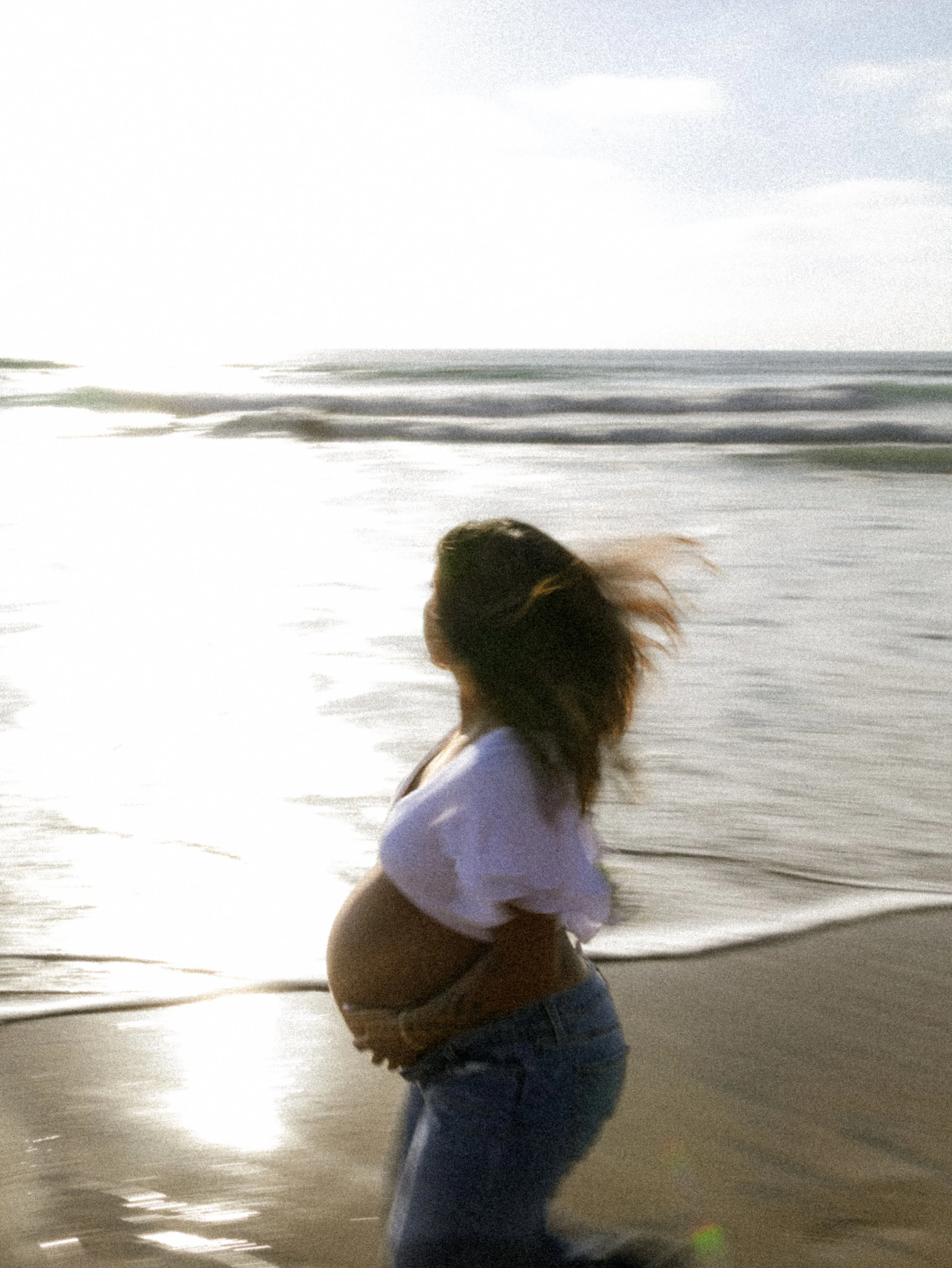 A woman with long hair in a white shirt and jeans is sitting on the beach, holding her knees, with the ocean and sunset in the background.
